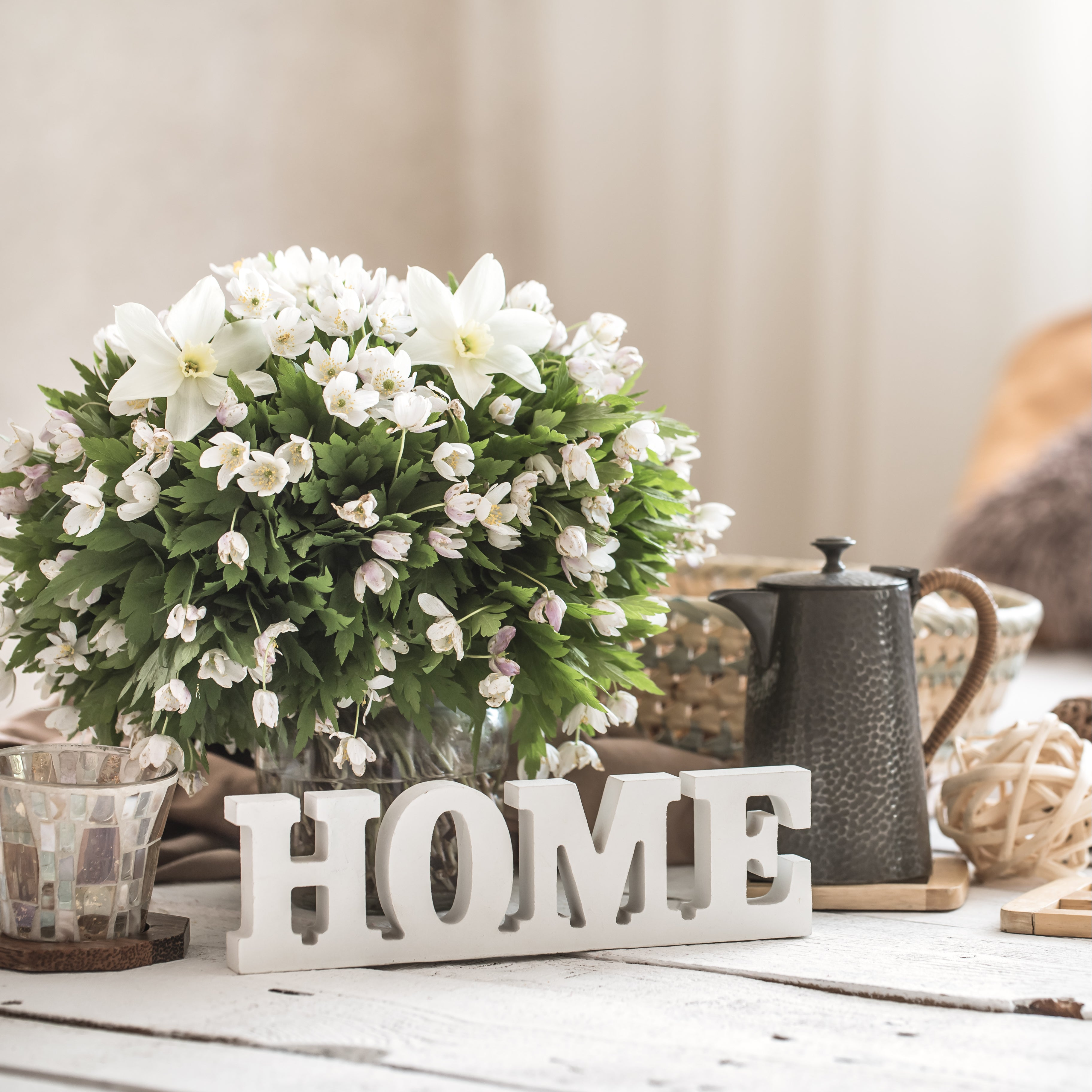 A "HOME" sign made of white-washed wooden letters atop a coffee table with a bouquet of flowers and home goods in neutral colors, with a window with translucent curtains in the background.