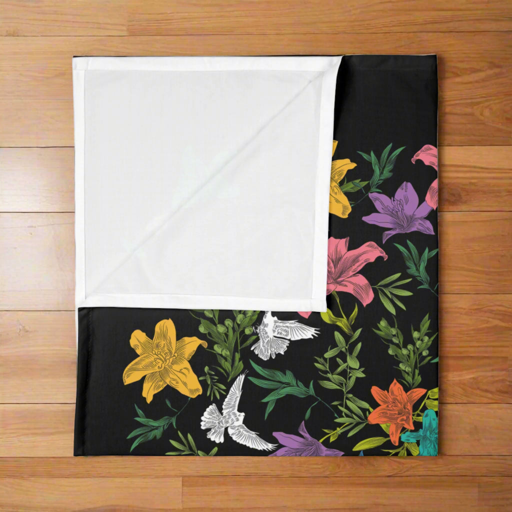 A folded, black knit, baby swaddle blanket with multi-colored, all-over print of doves, flowers, and olive branches, revealing a folded corner that displays the white underside.