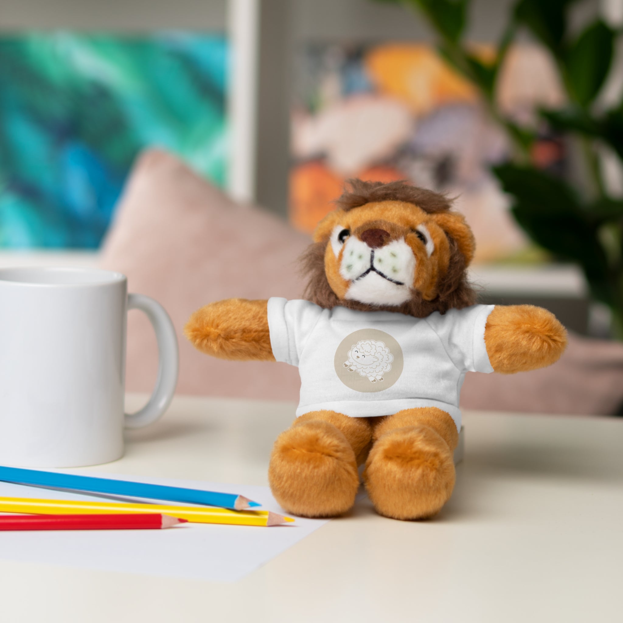 Plush lion toy wearing a white shirt with a lamb graphic on a table with stationery items.