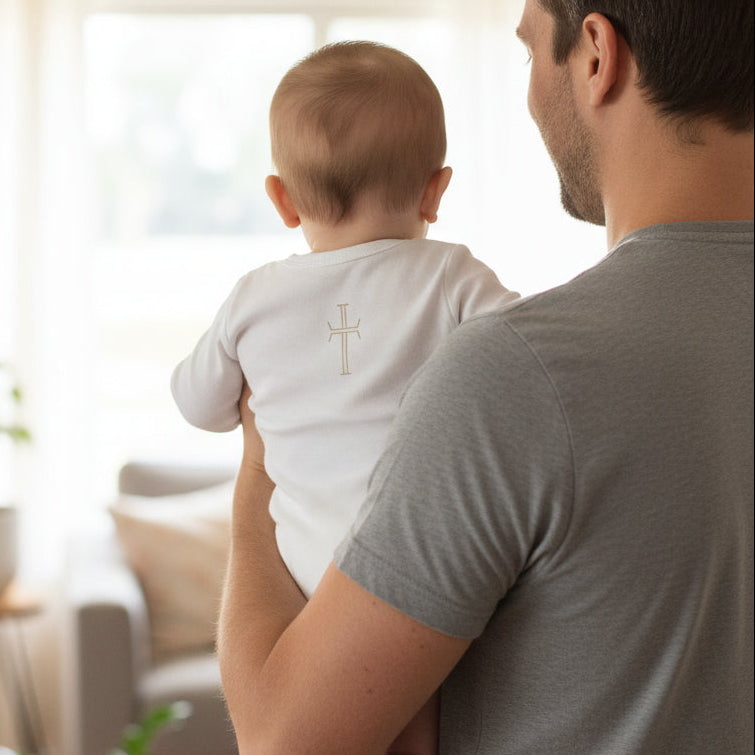 The back of infant in long-sleeve onesie with a cross on the back being held by father in a living room setting.