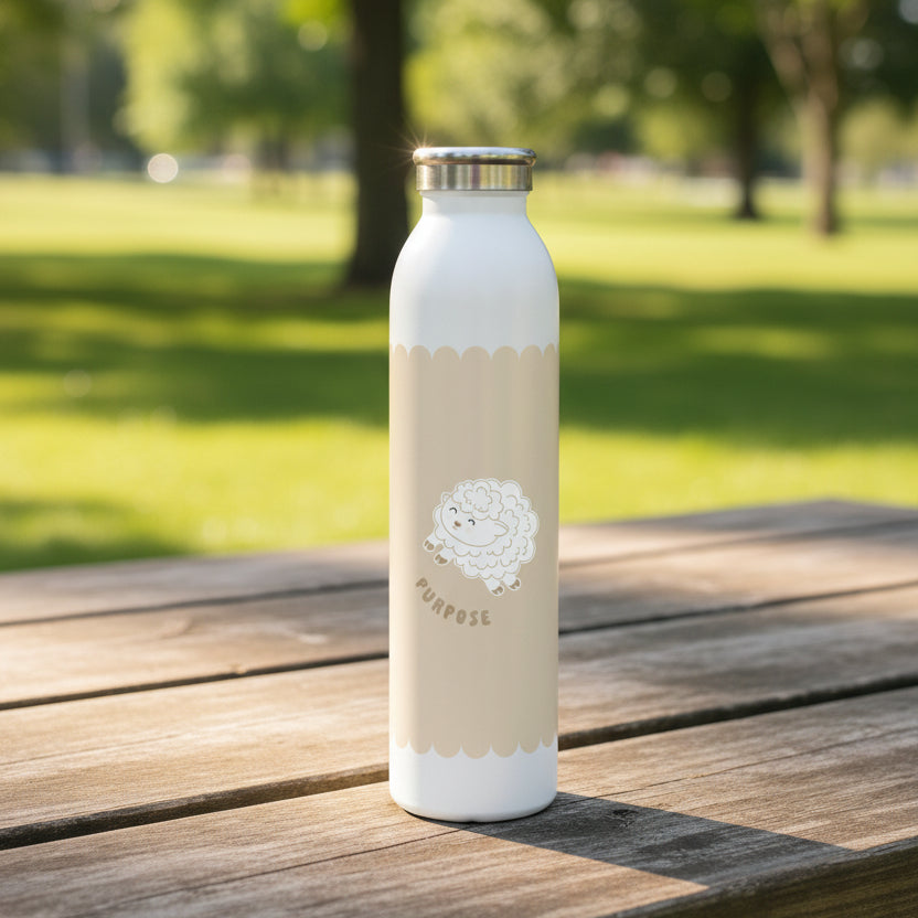 A white insulated water bottle with a silver metal cap, featuring a beige scalloped background around the middle with a cute white lamb illustration and the word 'PURPOSE' printed below it on a picnic table in a park.