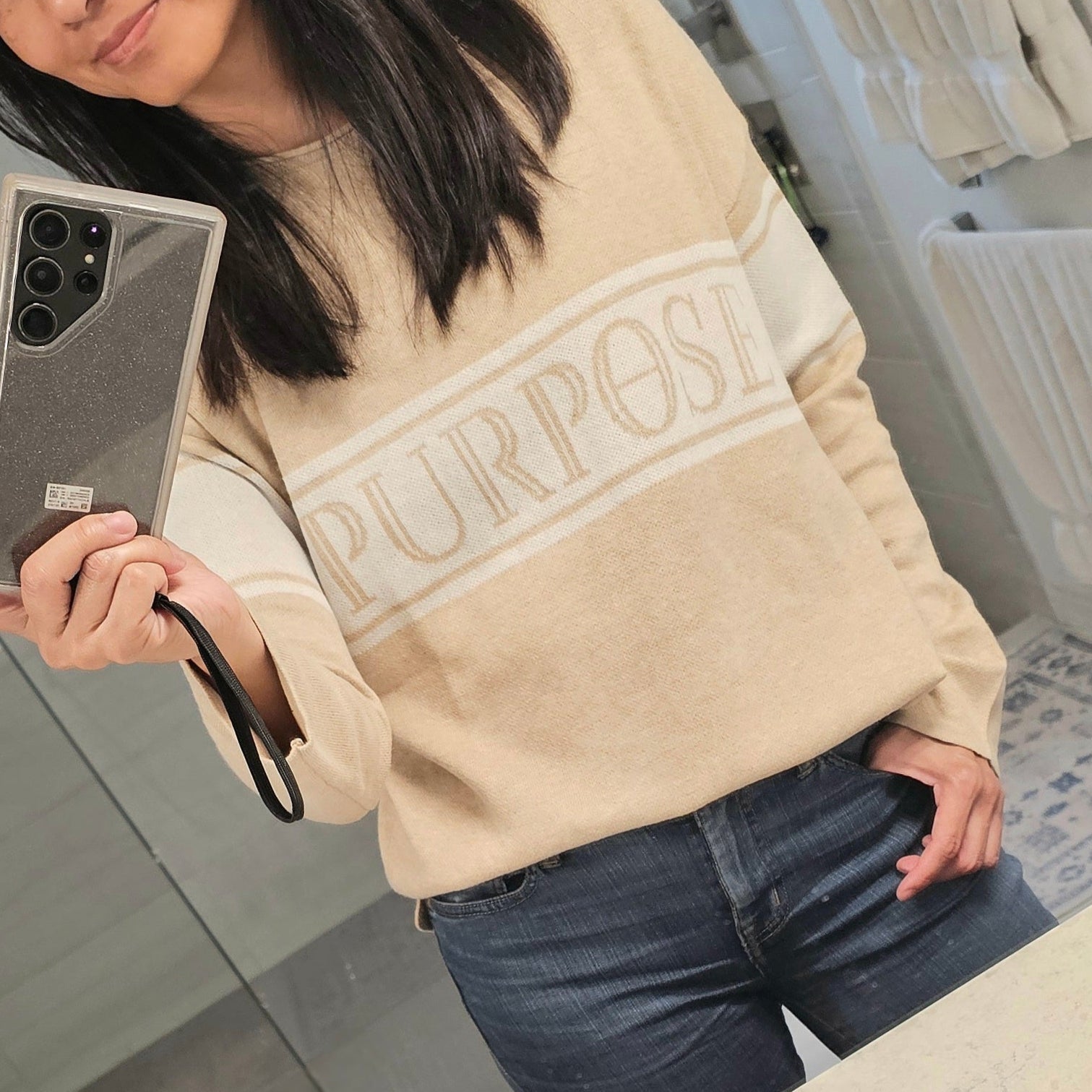 Person wearing a beige sweatshirt with 'PURPOSE' text, holding a phone in a bathroom.