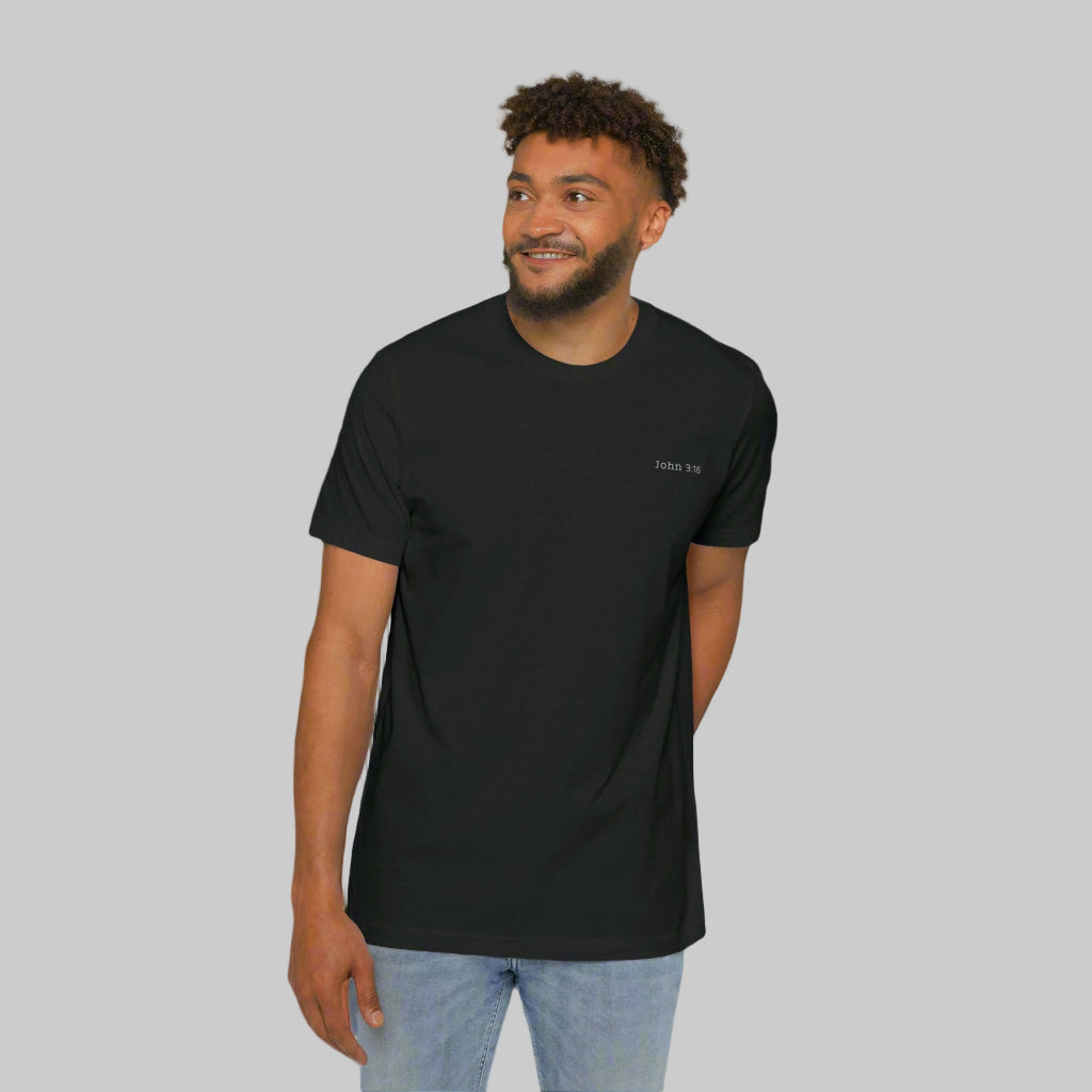 The front side of a male model wearing blue jeans and a black tee shirt with the bible verse John 3:16 written in white on the left chest.