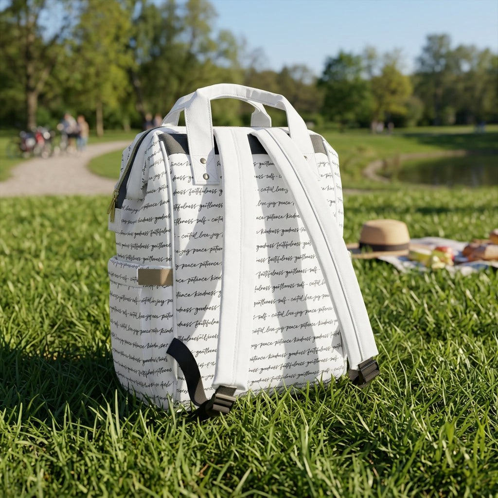 The back side of a white backpack with an all-over black script print featuring the Fruits of the Spirit with two padded shoulder straps, beige side accents, and a white top handle, shown on the grass in a park setting next to a picnic on a picnic blanket.