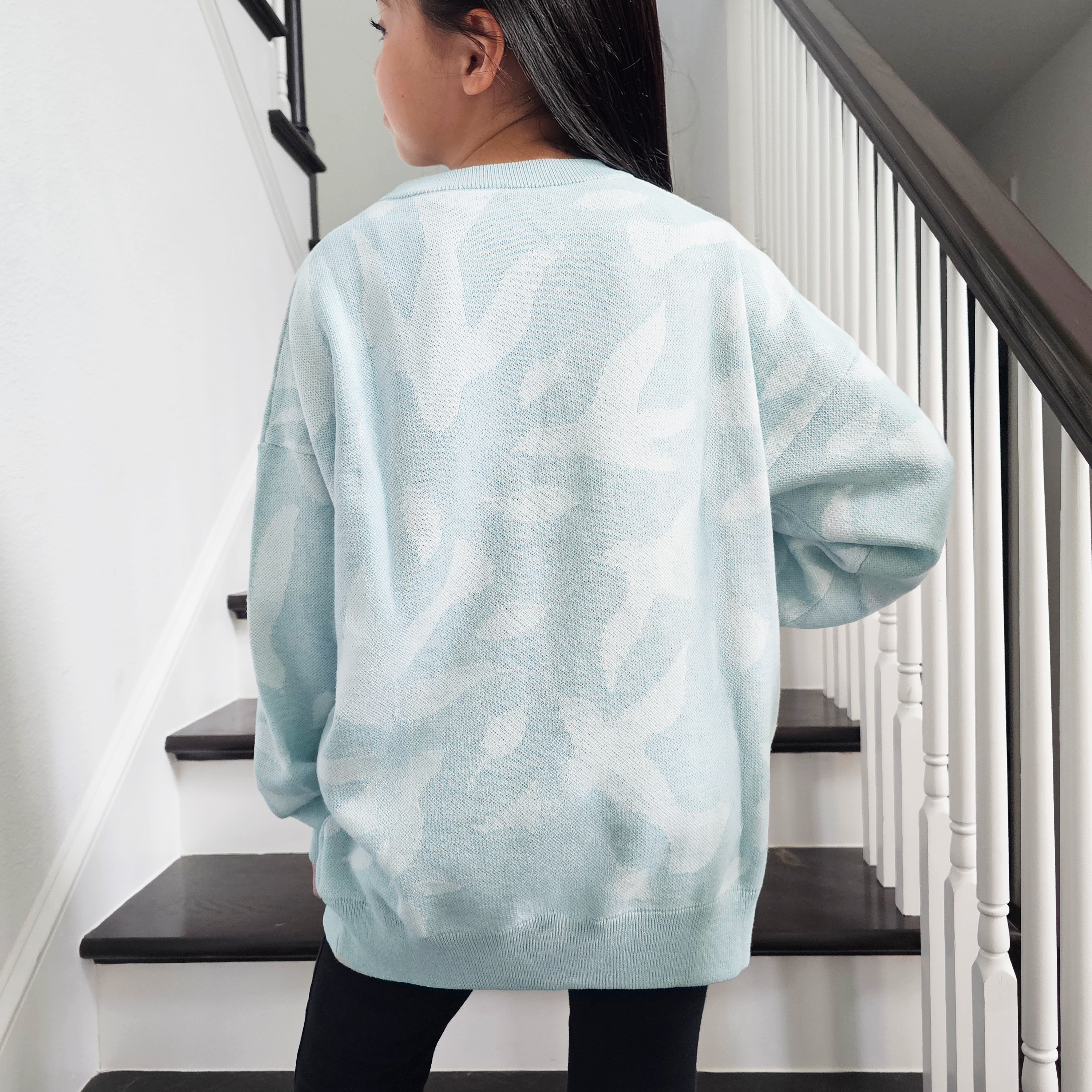 Young girl wearing a light blue knit sweater with white dove and leaf pattern standing on a staircase.