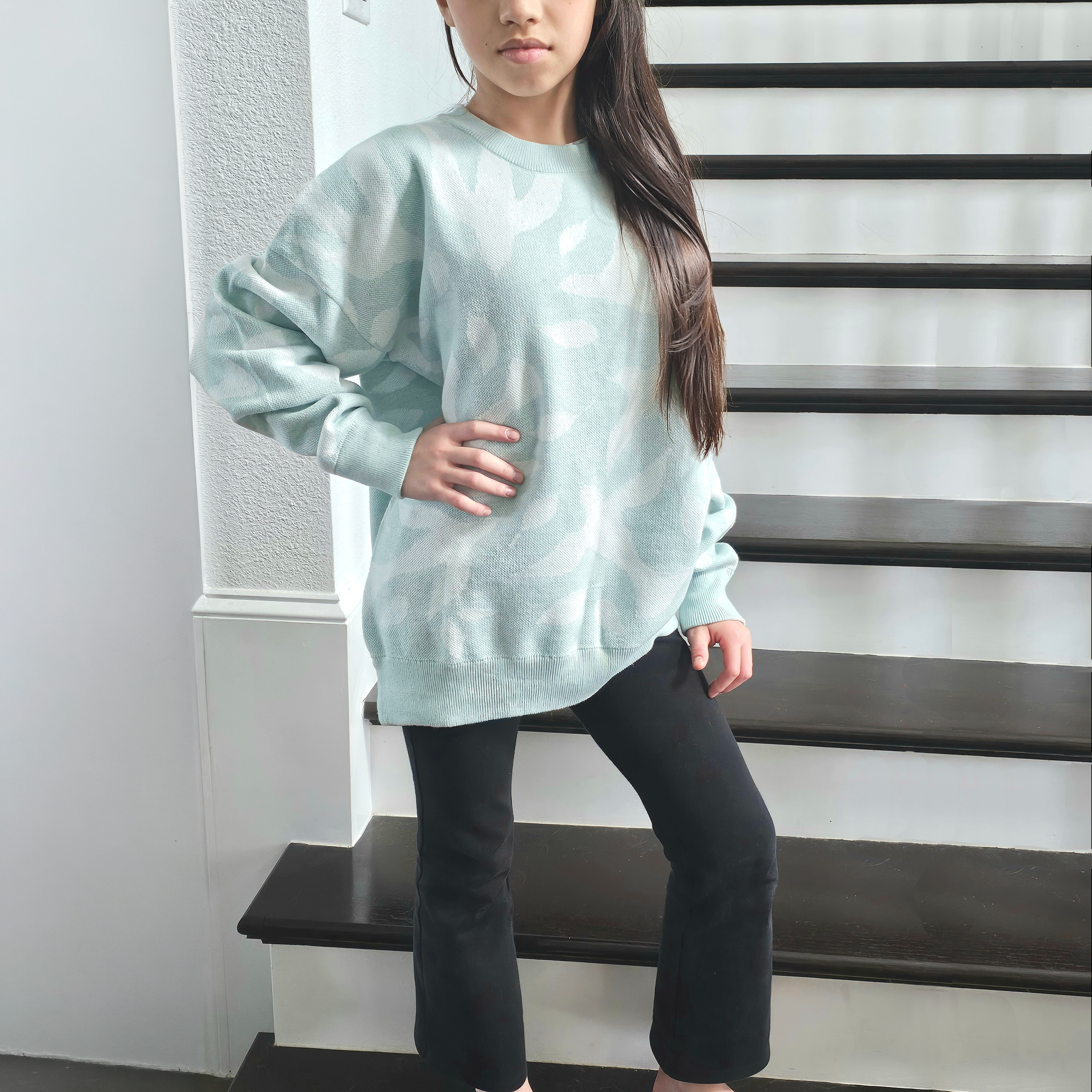 Young girl wearing a light blue sweater with white dove and leaf pattern and black pants standing on stairs.