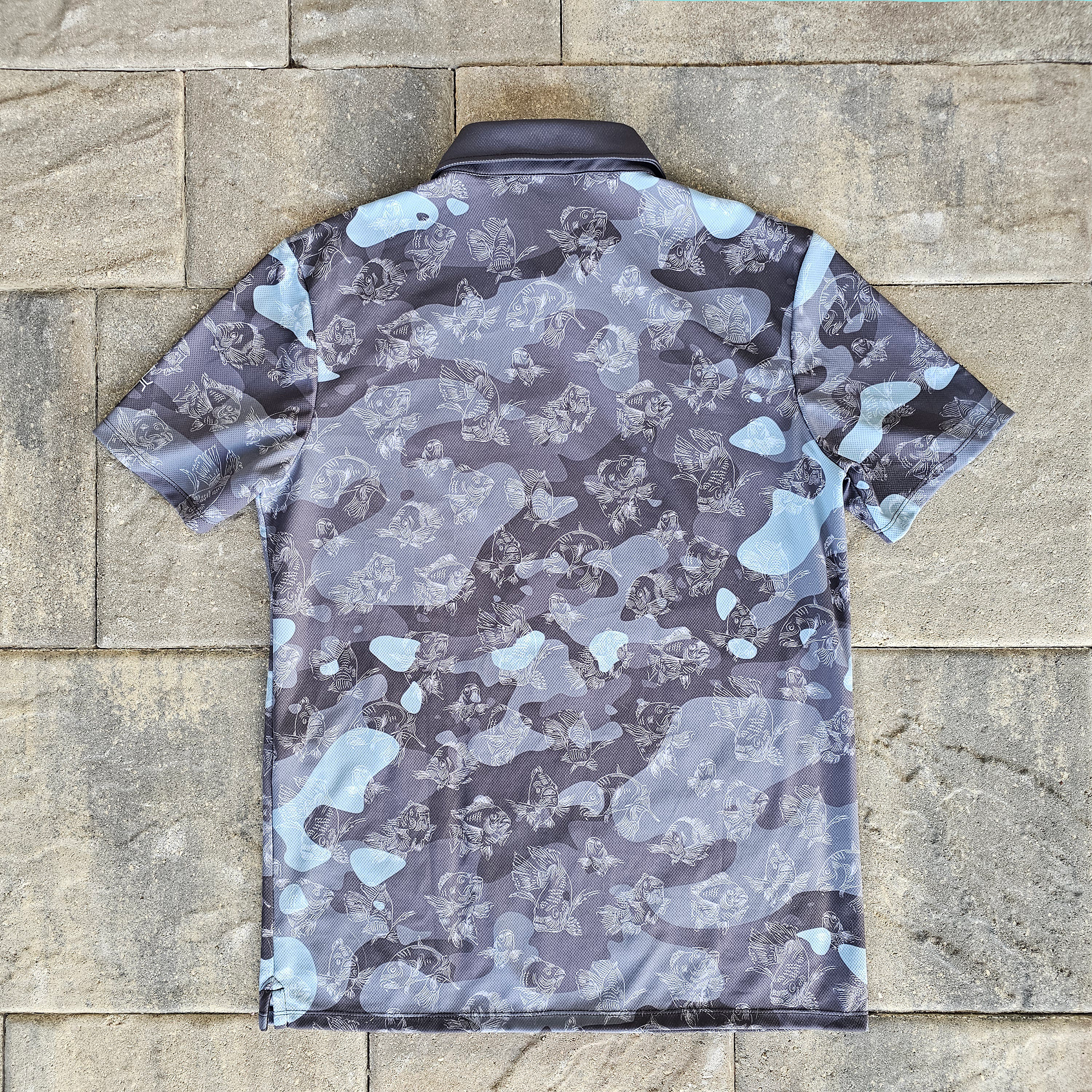 The back of a short-sleeve polo shirt with a gray, black, and light blue abstract camouflage pattern, laid flat on a stone tile background.