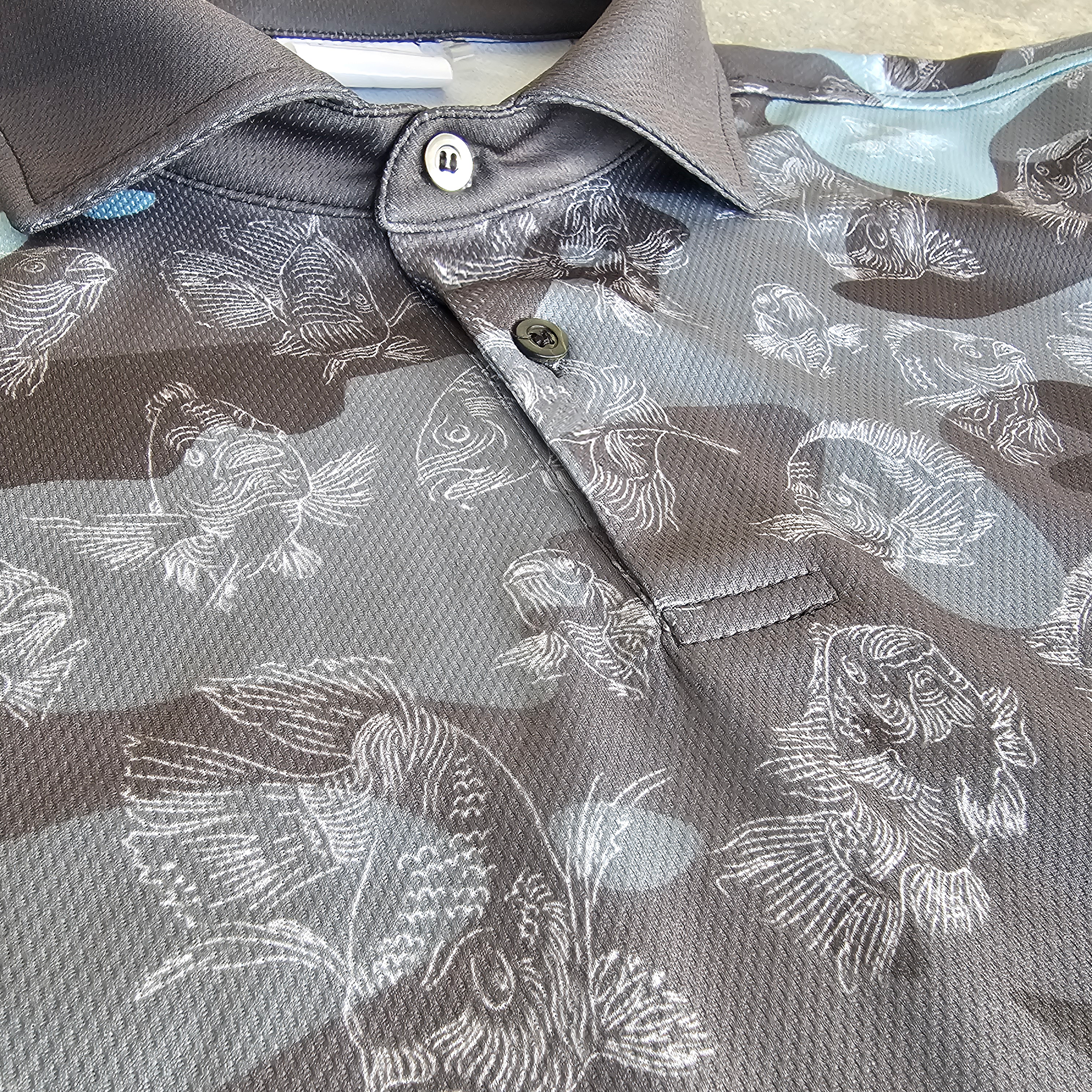 Close-up of a collared short-sleeve polo shirt with a gray, blue, and black camouflage pattern overlaid with white carp fish illustrations, featuring a two-button placket.