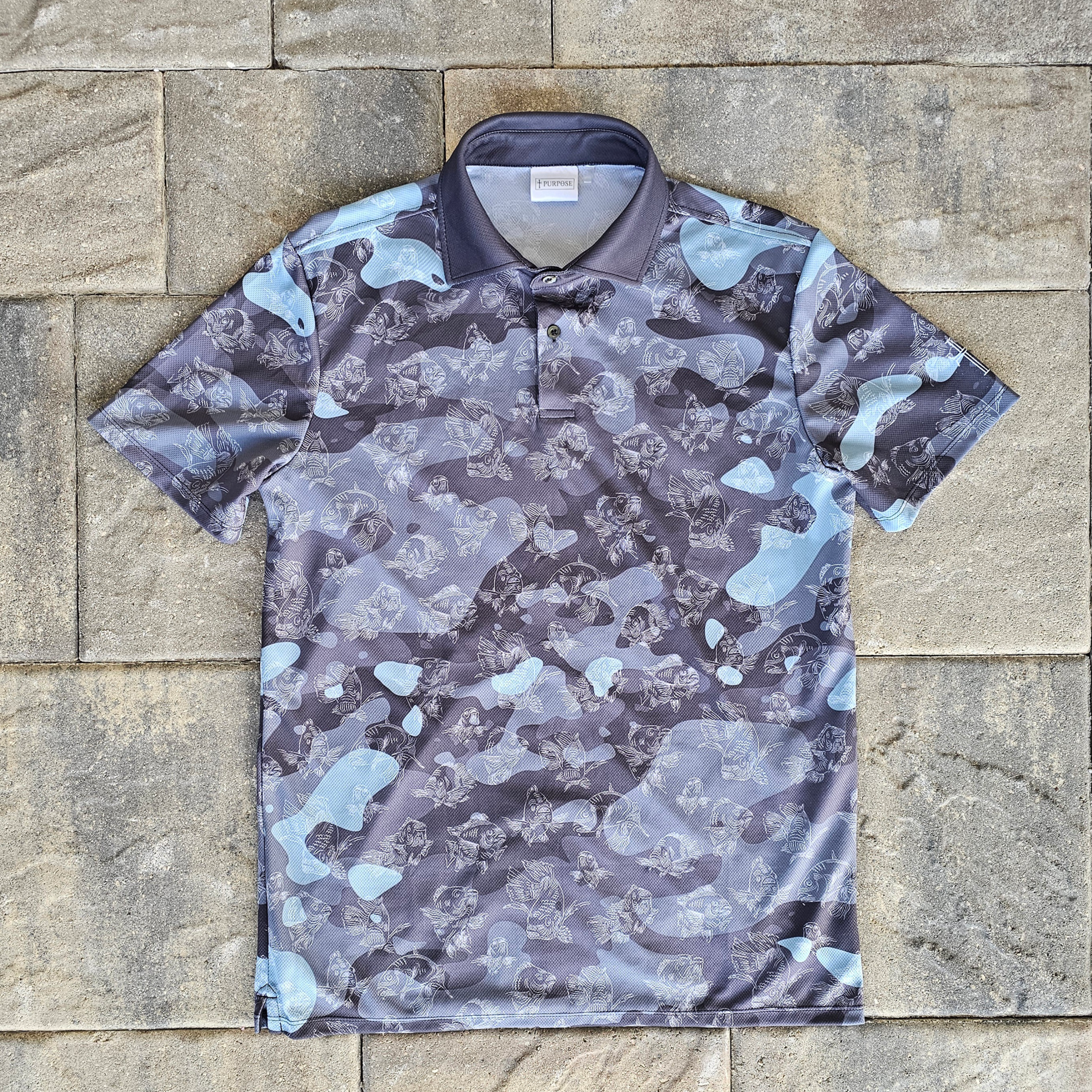 A short-sleeve polo shirt with a gray, black, and light blue camouflage pattern, laid flat on a stone tile background.