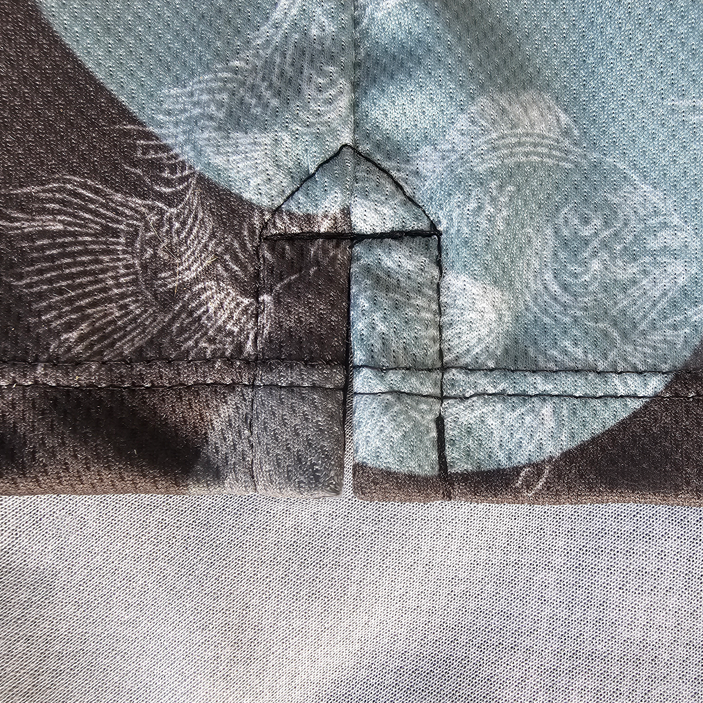 Close-up of a garment construction detail showing the front of a reinforced placket hem with a zig-zag fold and whipstitch, in a black, gray, and light blue camo fabric white carp graphic pattern.