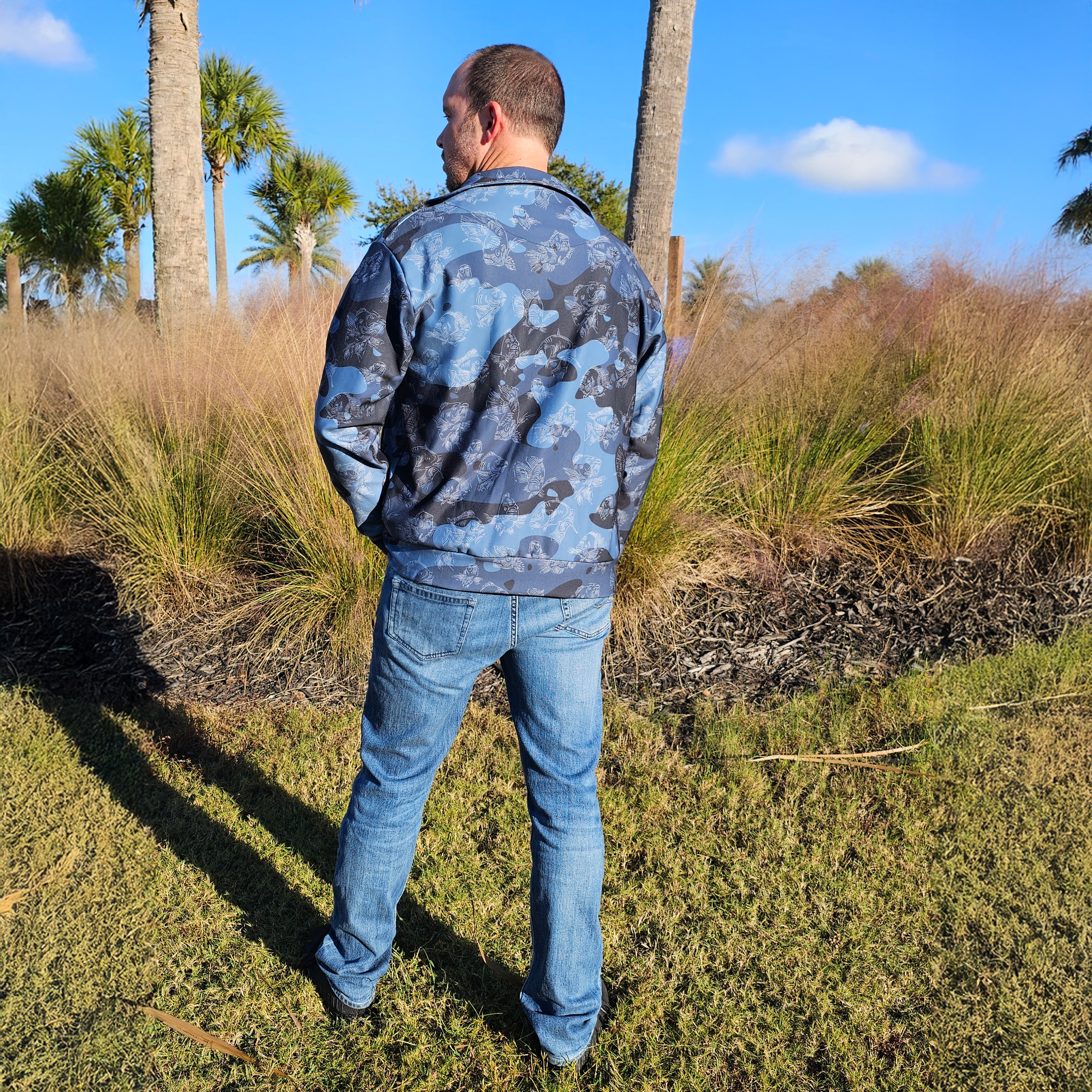 Man wearing a camouflage jacket and jeans standing in a grassy area with palm trees.