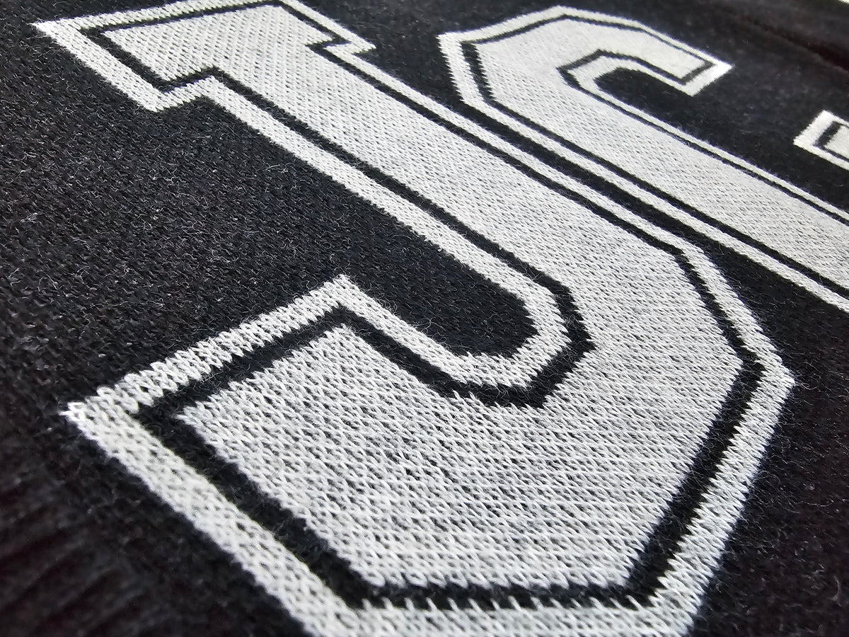 Close-up of a black knit fabric with a large white outlined letters 'JC'' knitted across it.