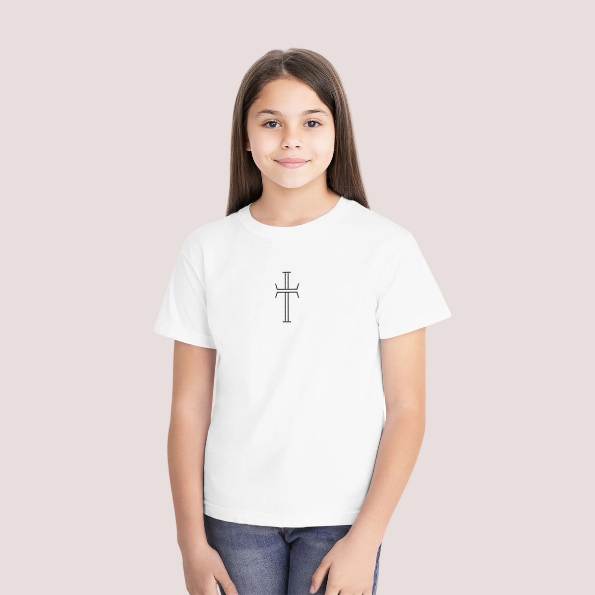 A young girl with long brown hair stands against a solid white background wearing a white short-sleeve crewneck t-shirt with a small black cross printed on the center chest, paired with blue jeans.