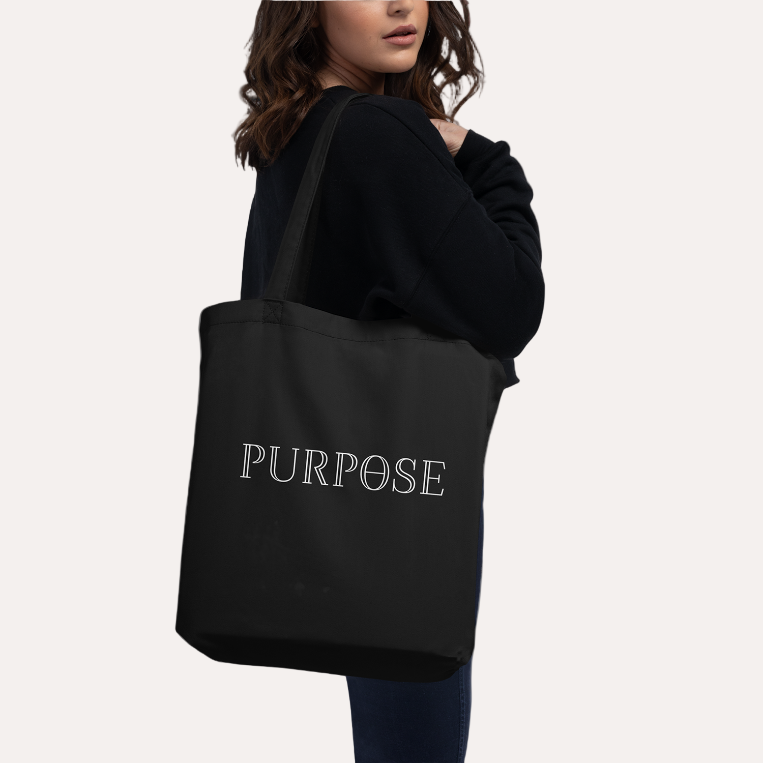A female model displays the back of a black tote bag over her shoulder. It has two long handles and the white Purpose worded logo printed on the back of the bag.