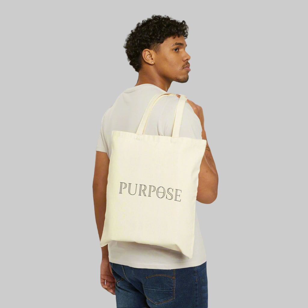 A male model in a tee shirt and jeans is slinging a tote bag with long double handles over the back of his shoulder. The natural canvas bag has the black Purpose word logo on the back of the bag.
