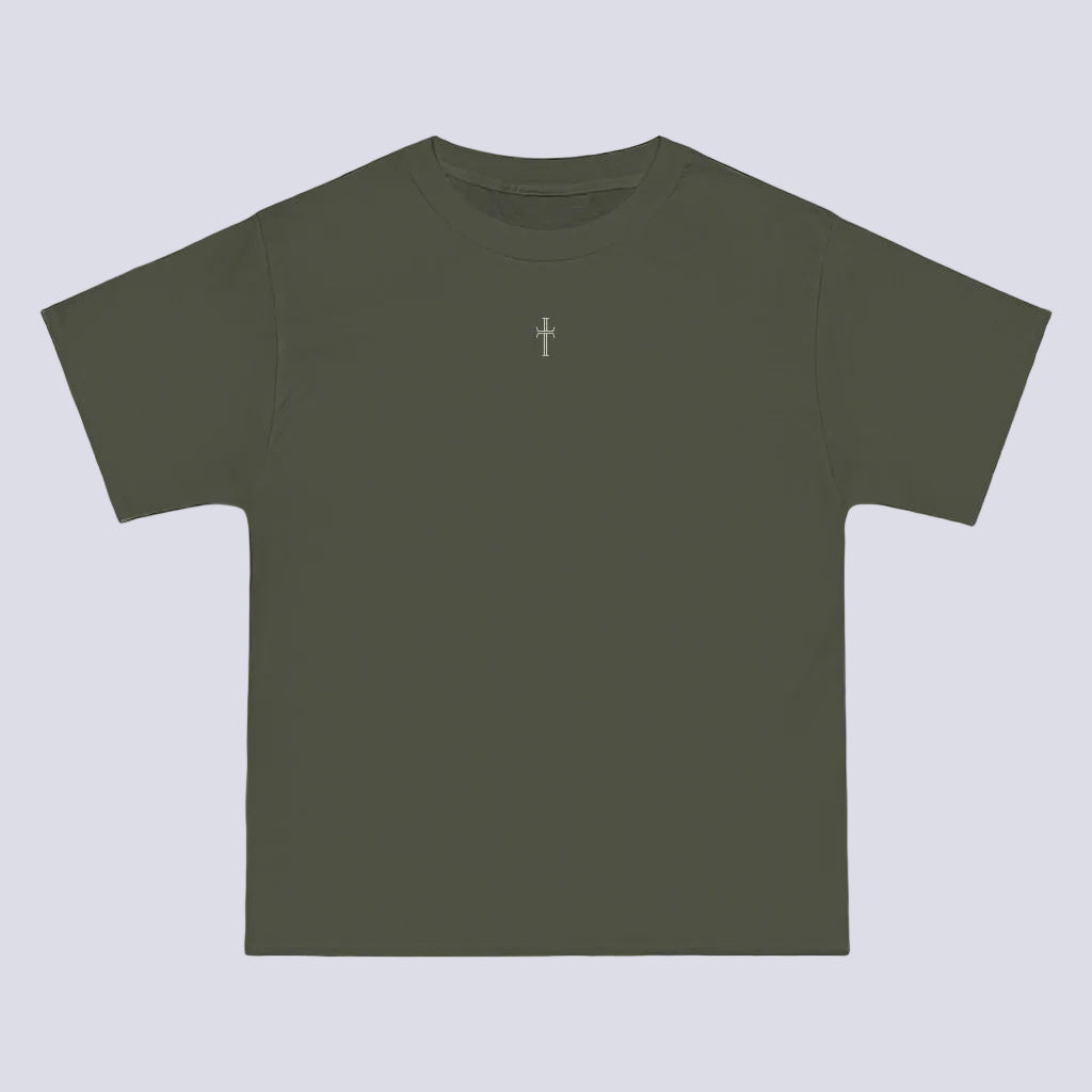 An army green crewneck tee shirt displays the front of the garment as it lays against a brick-patterned wall. A small white Purpose cross logo is printed at the top center.