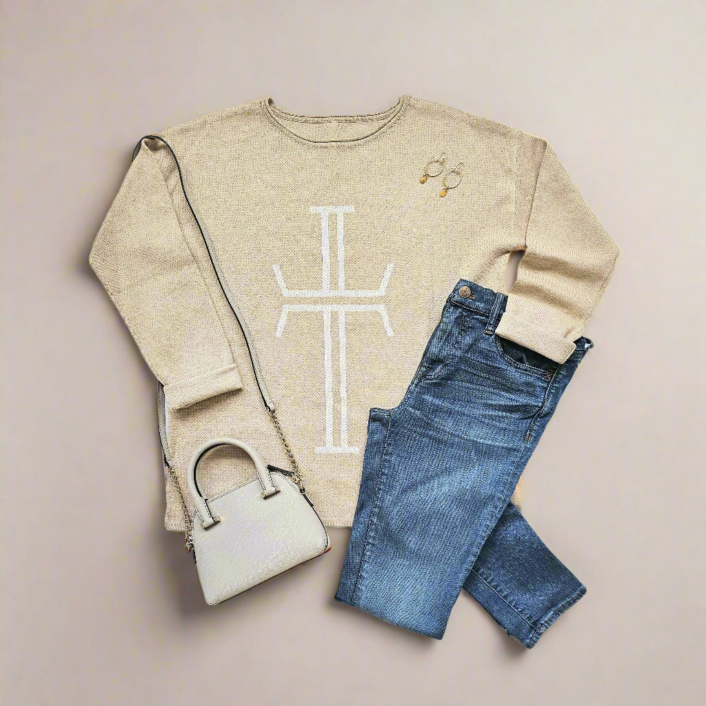 A beige sweater with a straight hem and plain cuffs and neckline is displayed against a white background. A bold, white, stylized crucifix is knitted along the entire front of the sweater. A cream-colored mini purse with long shoulder strap, blue denim jeans, and a pair of dangly earrings are displayed around the sweater. Christian sweater.