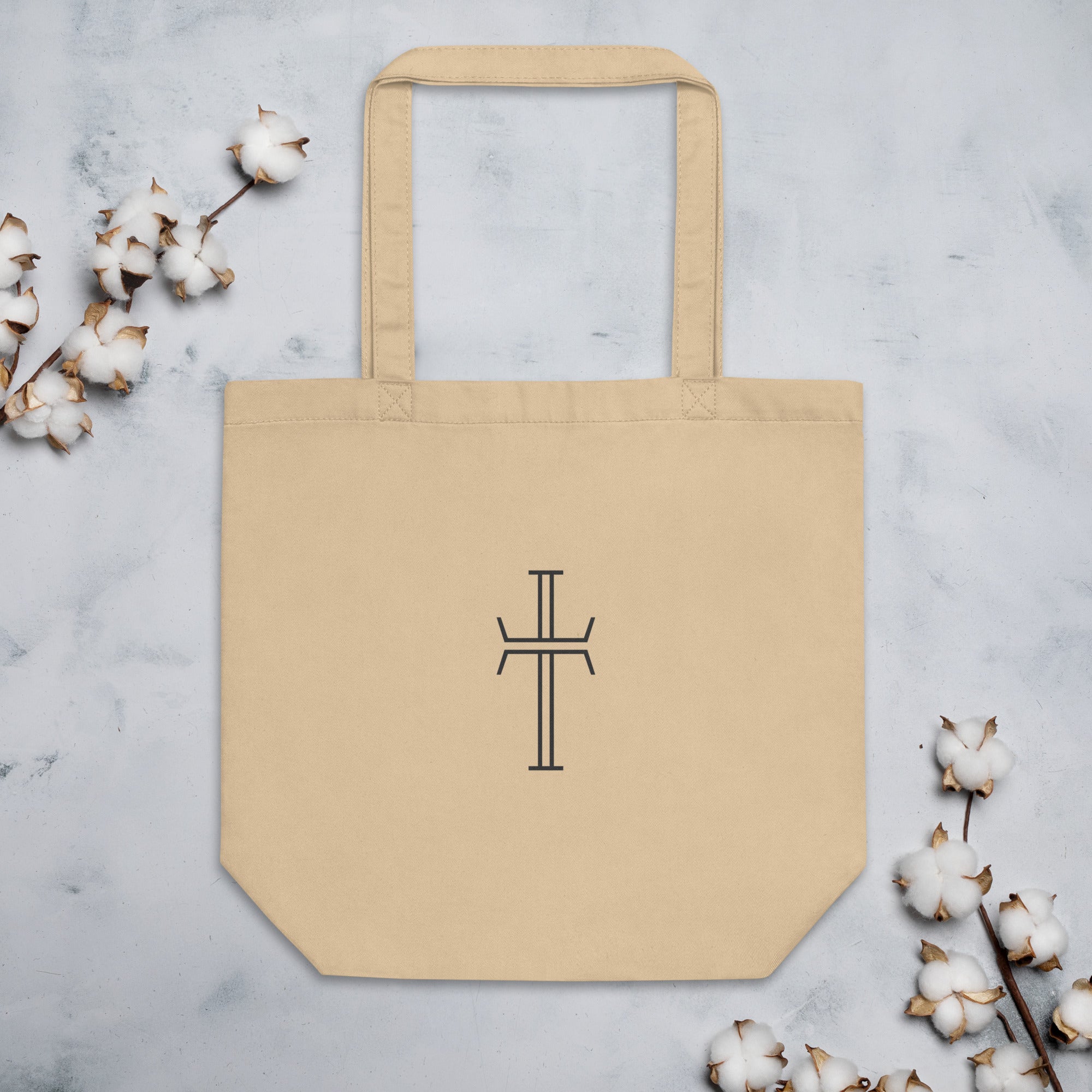 Natural-colored cotton canvas tote bag with shoulder straps and a centered black stylized cross on the front.