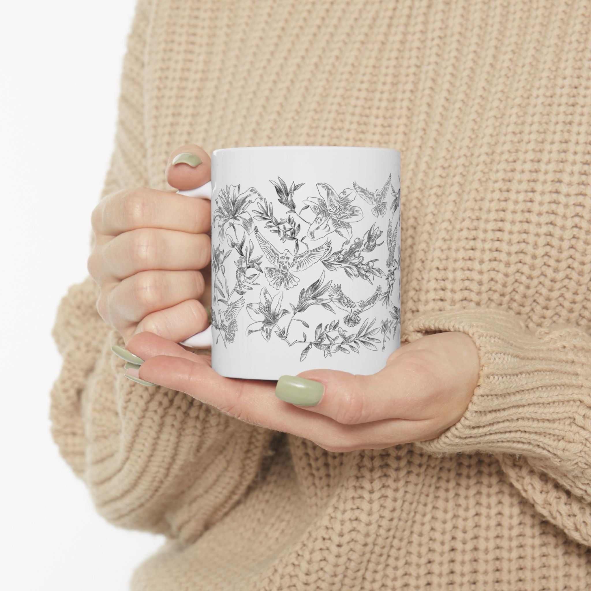 Woman holding a white mug with black floral and dove design, wearing a beige sweater.