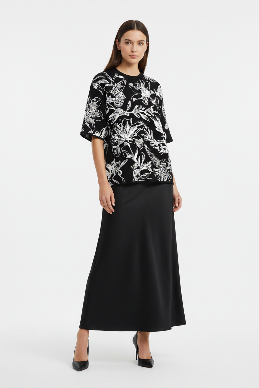 Full-length front view of young woman wearing black short-sleeve sweater with white knitted floral and dove pattern, long black skirt, and black pumps.