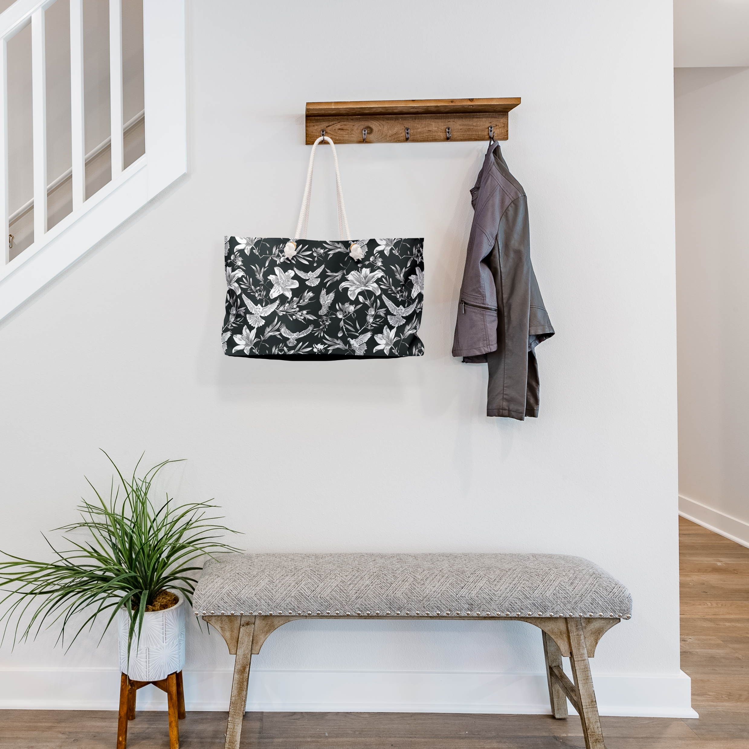 A large-capacity, black, weekender bag with thick rope handles, hangs from a jacket rack in the entryway of a transitionally-styled house of neutral colors. The bag has a bold white pattern of doves, lilies, and olive branches in an all-over printed pattern. Grommets to accommodate the rope handles are gold-colored.