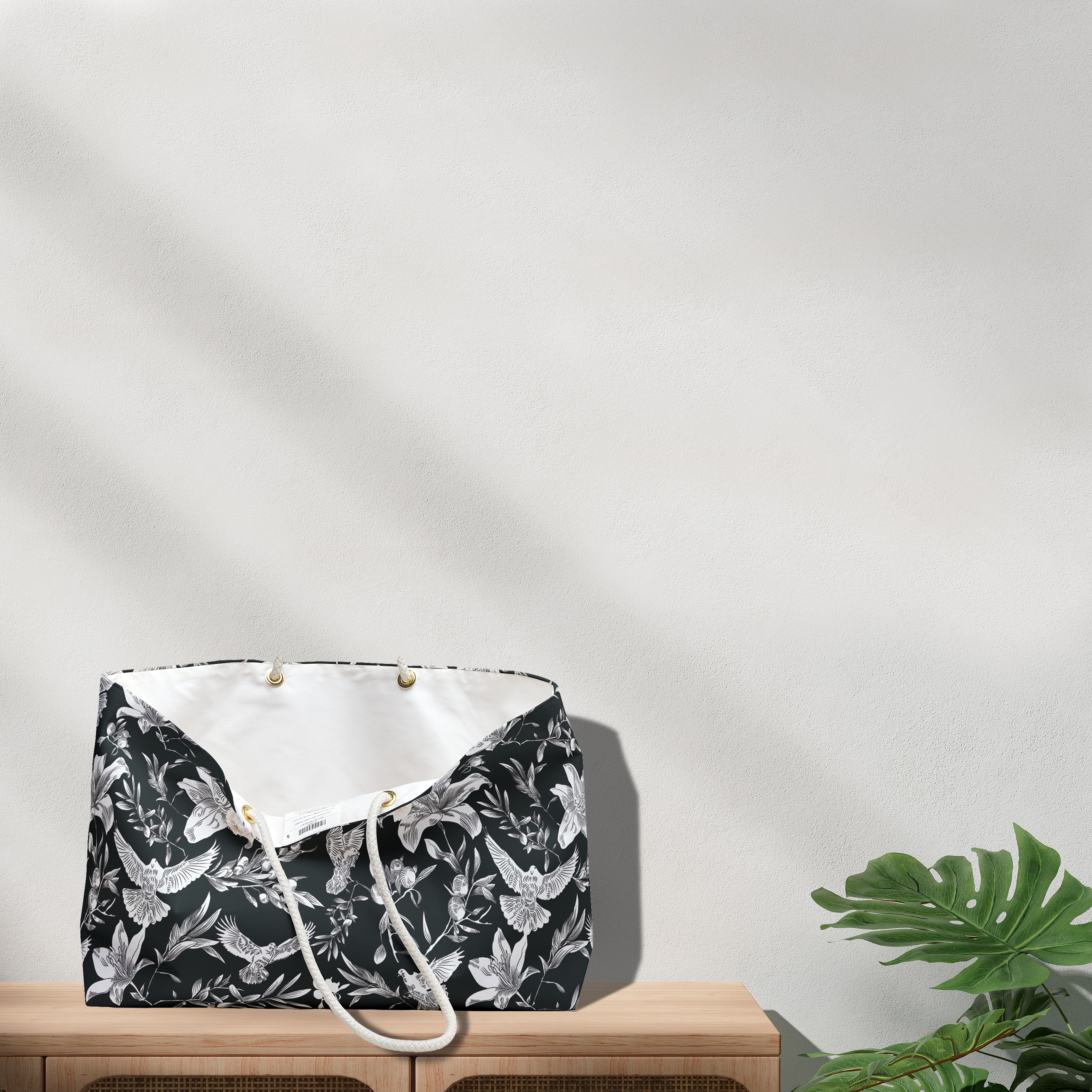 A large-capacity, black, weekender bag with white interior and thick rope handles stands open atop a light-colored wood console next to a green, large-leafed plant. The bag has a bold white pattern of doves, lilies, and olive branches in an all-over printed pattern. Grommets to accommodate the rope handles are gold-colored.