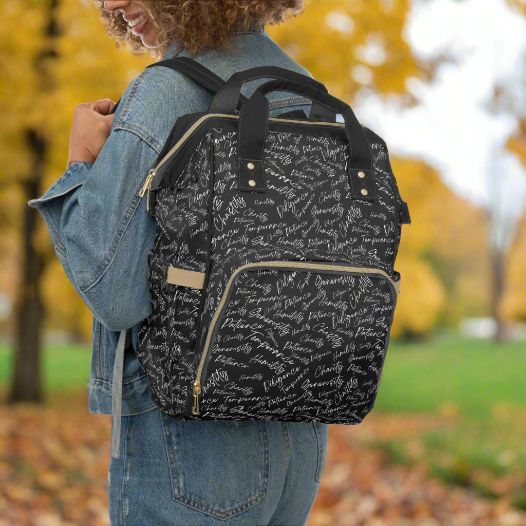 A person is shown from the back wearing a black and white diaper backpack with multiple compartments and adjustable shoulder straps.