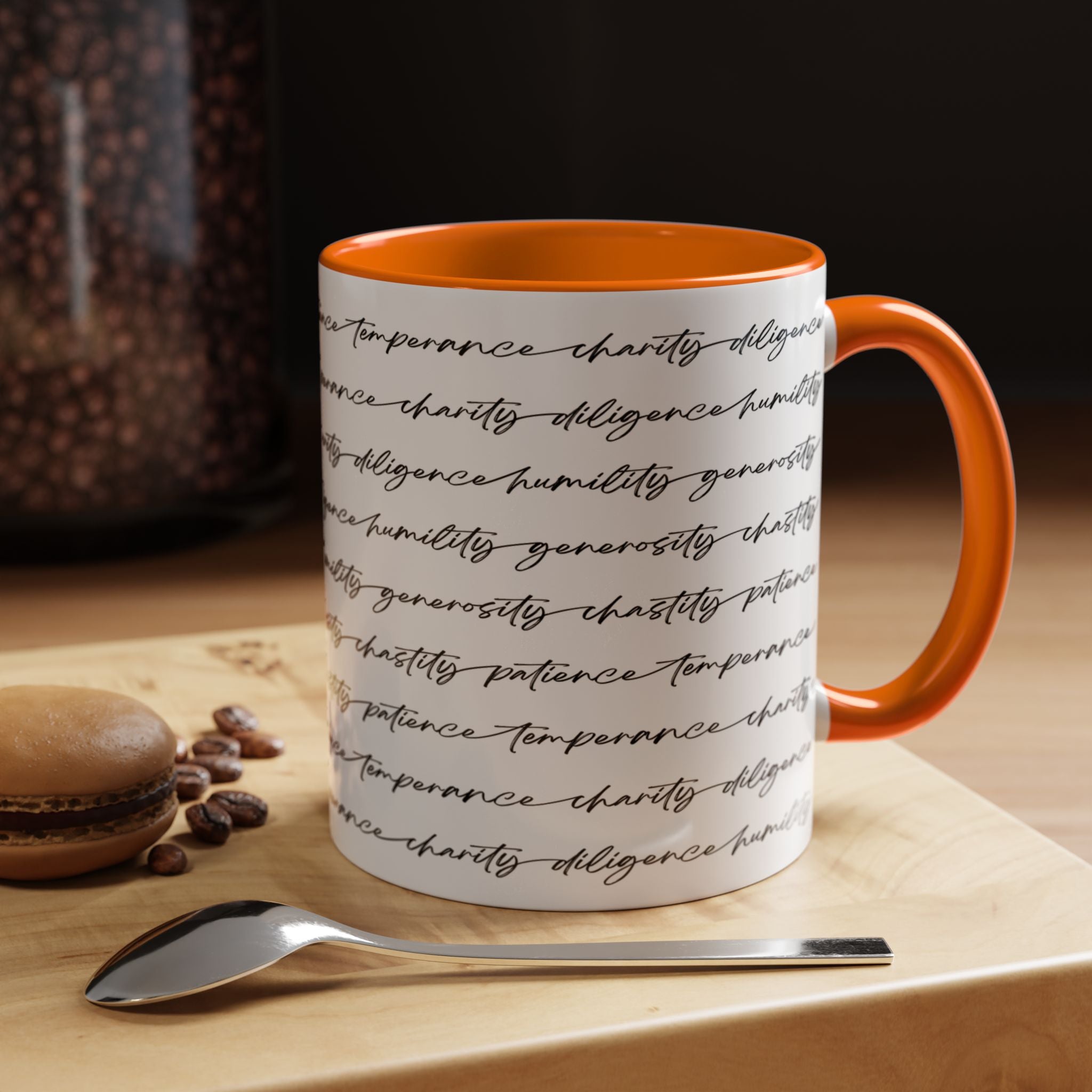 White mug with the seven virtues scripted along the outside, with orange interior and handle on a wooden surface with coffee beans and a spoon.