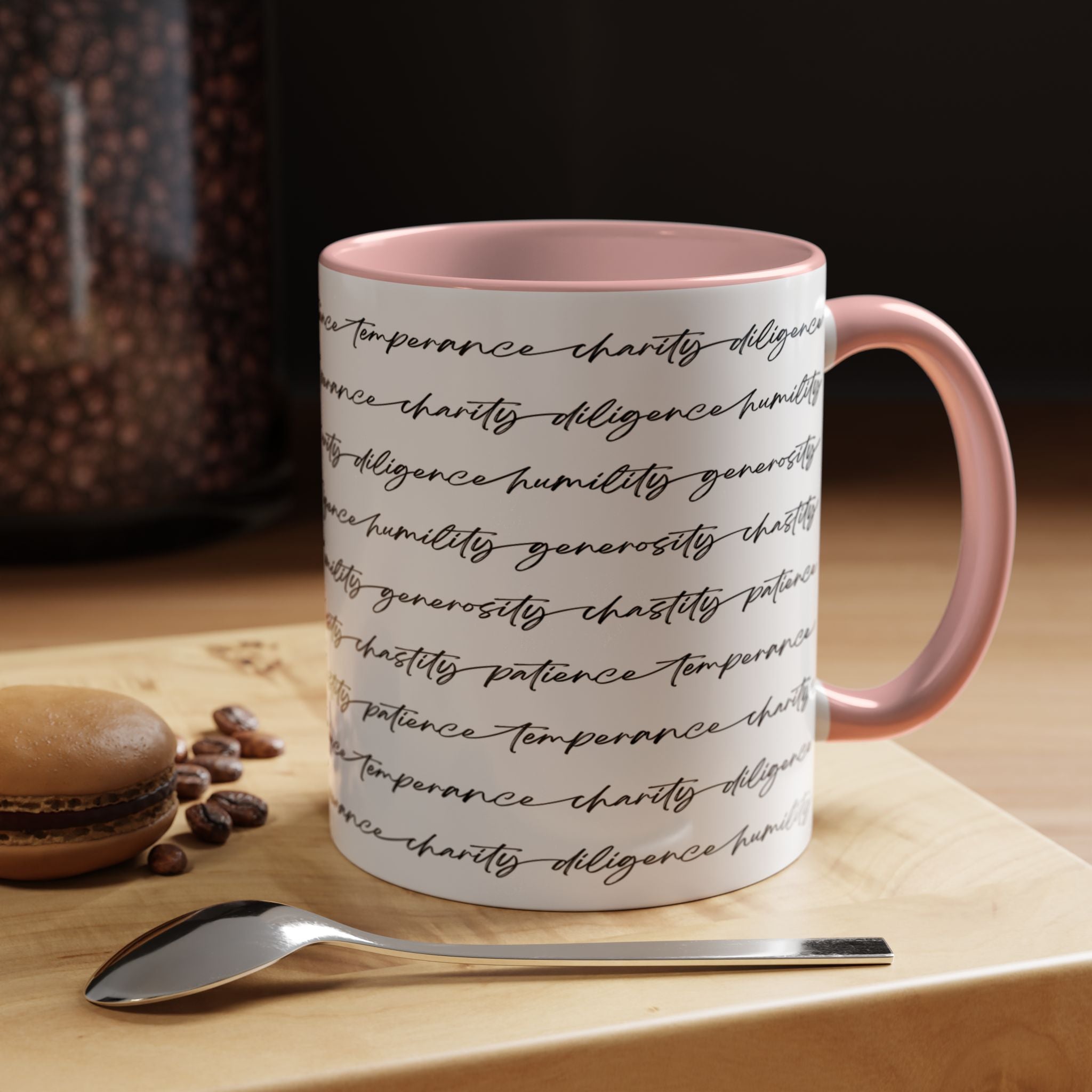 White mug with the seven virtues scripted along the outside, with pink interior and handle on a wooden surface with coffee beans and a spoon.