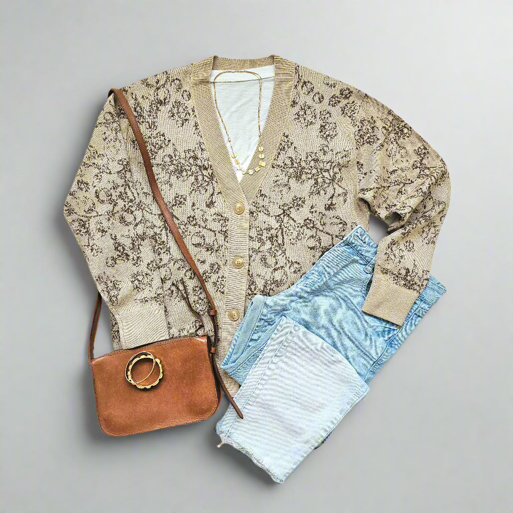 A beige knit, v-neck cardigan with large beige buttons is displayed against a white background. A brown floral pattern is knitted in an all-over pattern along the front. A pair of ombre-faded blue jeans, a small, camel colored shoulder bag, two gold-toned bangles and a gold-toned necklace are arranged around the cardigan. A white t-shirt peeks out from inside the sweater.