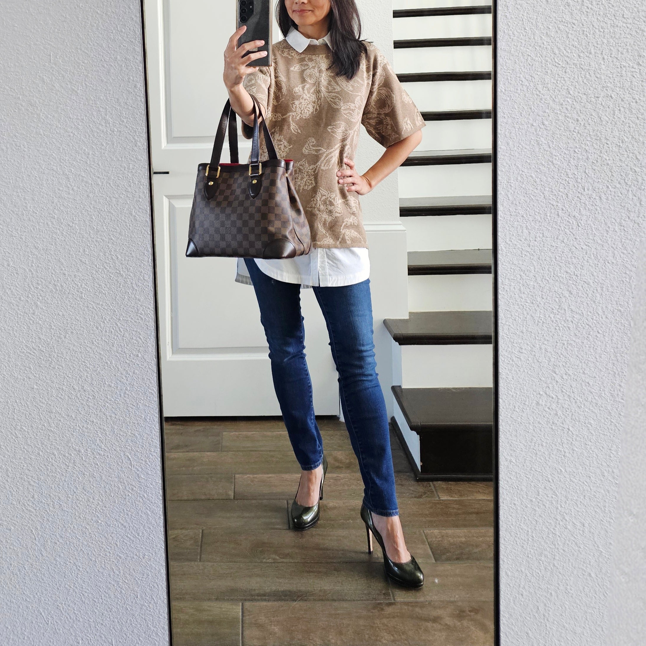Person wearing a patterned top, white shirt, blue jeans, and black heels, holding a brown handbag.