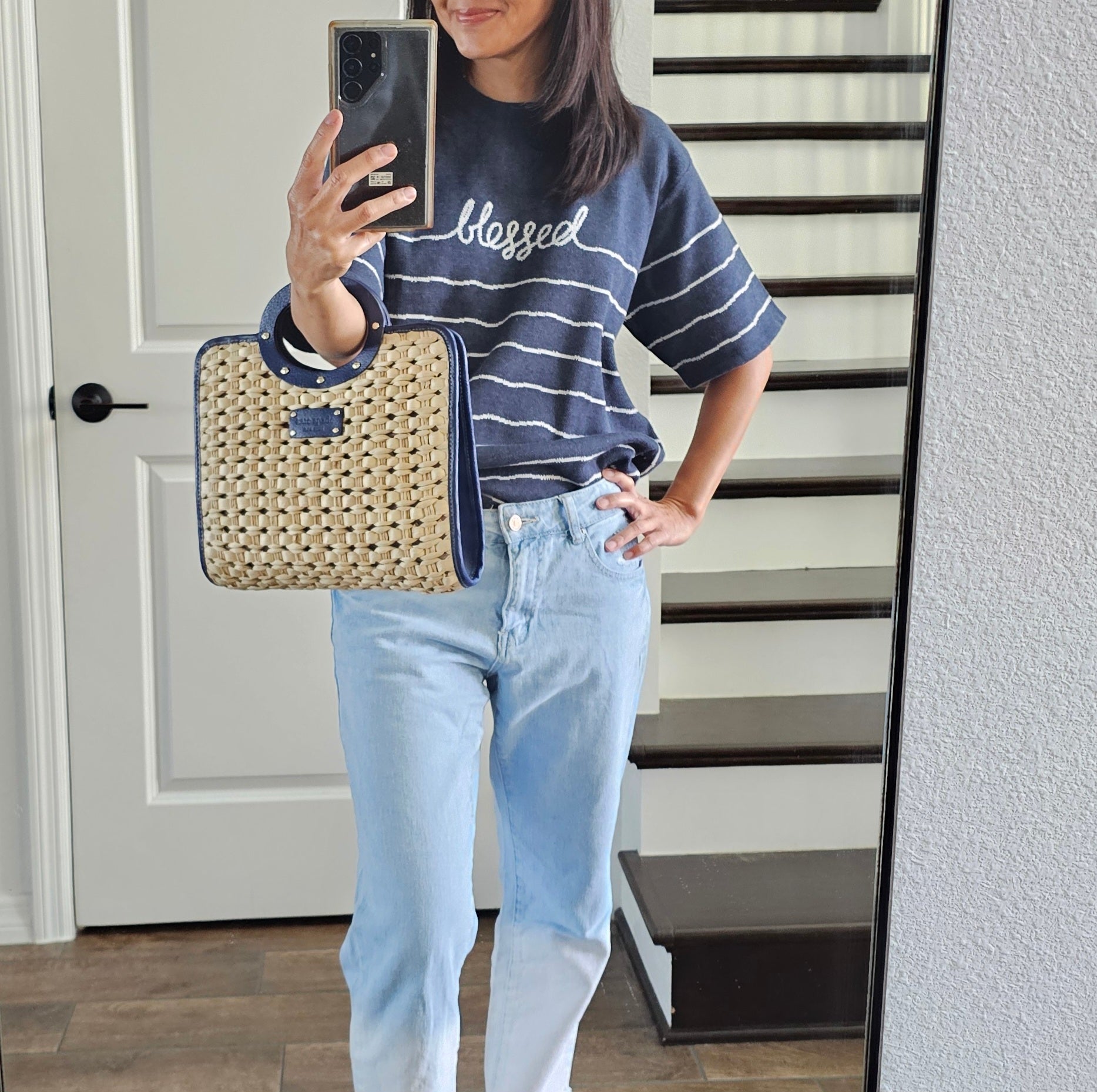 Person wearing a navy blue 'blessed' shirt and light blue jeans, holding a woven handbag.