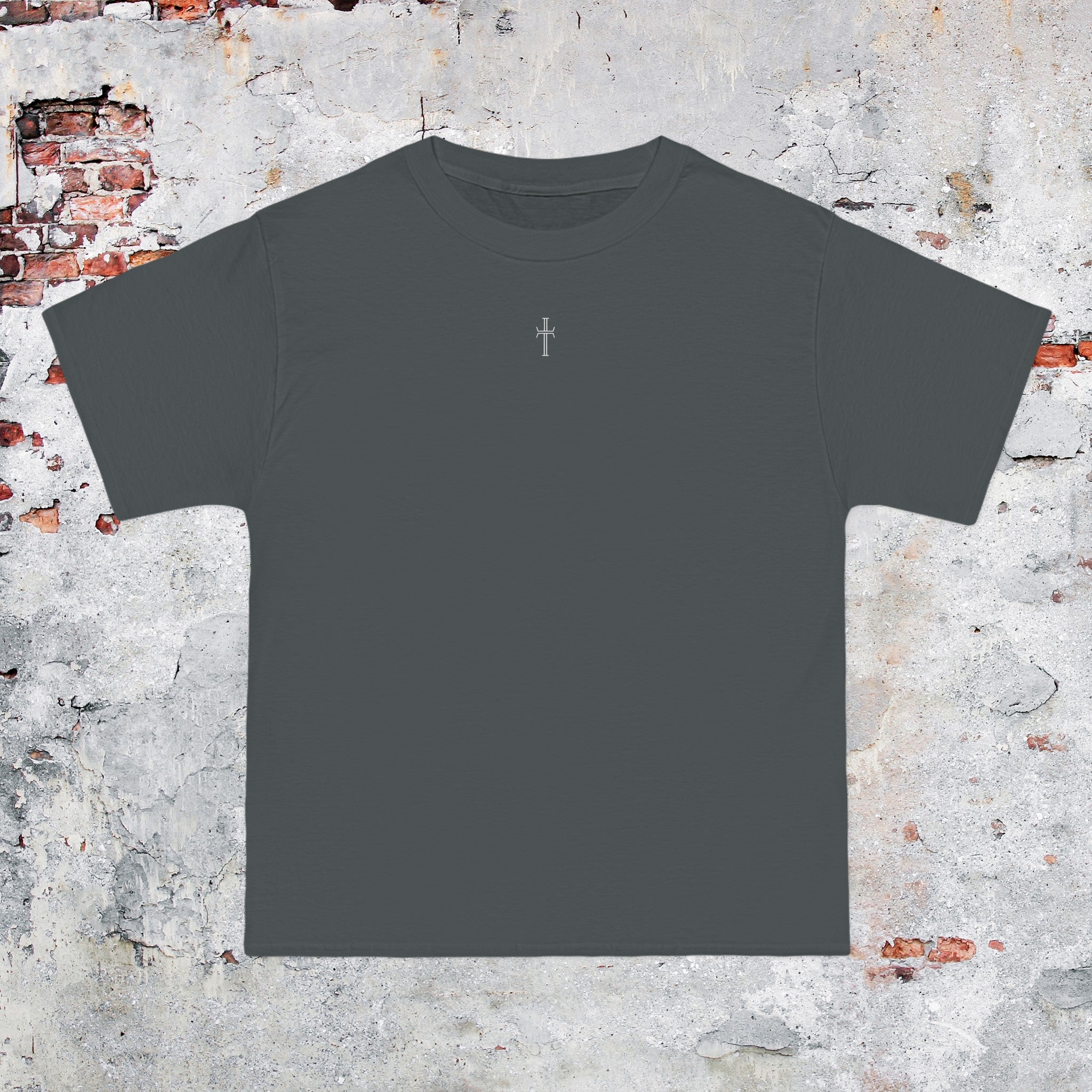 A gray crewneck tee shirt displays the front of the garment as it lays against a brick-patterned wall. A small white Purpose cross logo is printed at the top center. 