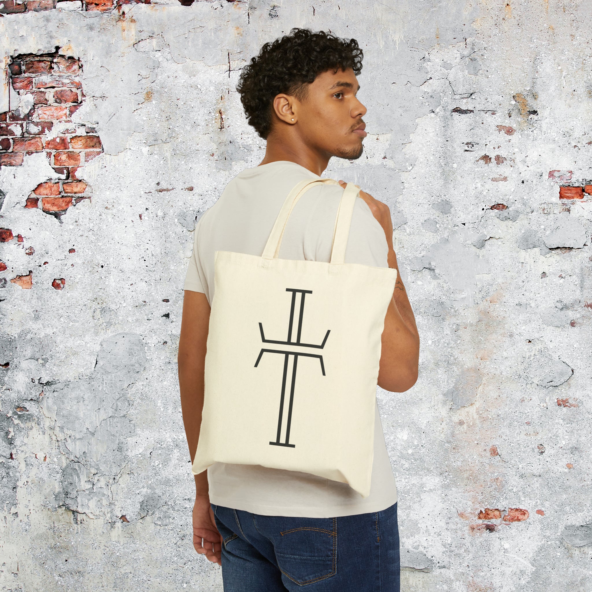 A male model in a tee shirt and jeans is slinging a tote bag with long double handles over the back of his shoulder. The natural canvas bag has a black Purpose cross logo on the front.