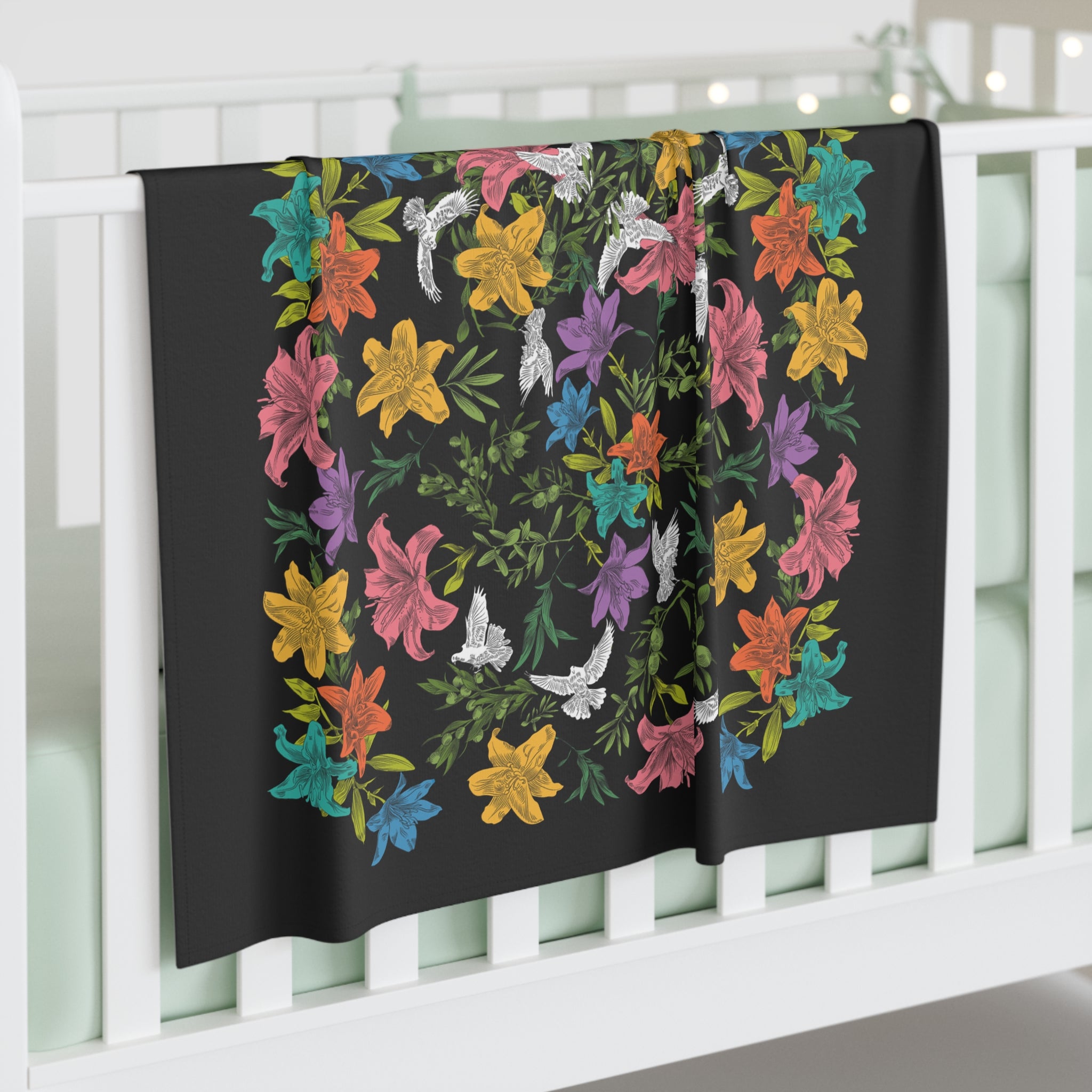 A black swaddle blanket draped over the side of a crib. The blanket has an all-over, multi-colored pattern of doves, flowers, and olive branches.