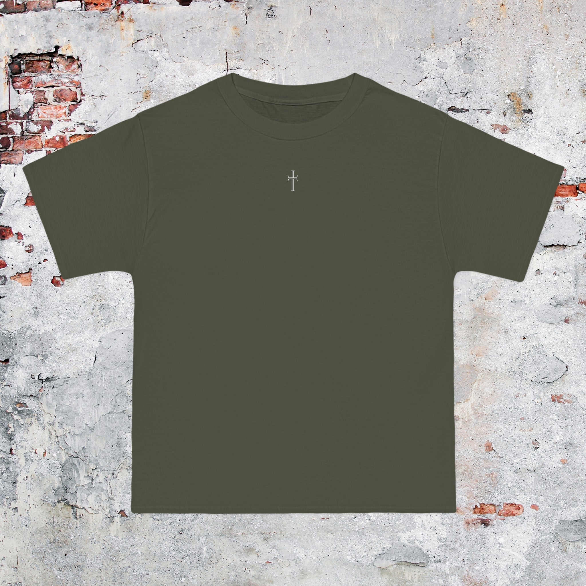 An army green crewneck tee shirt displays the front of the garment as it lays against a brick-patterned wall. A small white Purpose cross logo is printed at the top center. 