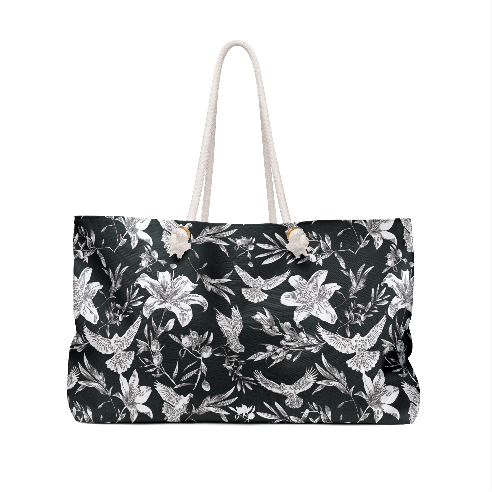 A large-capacity, black, weekender tote with a white, all-over pattern of doves, lilies, and olive branches with thick rope handles and gold-colored grommets stands against a white background.