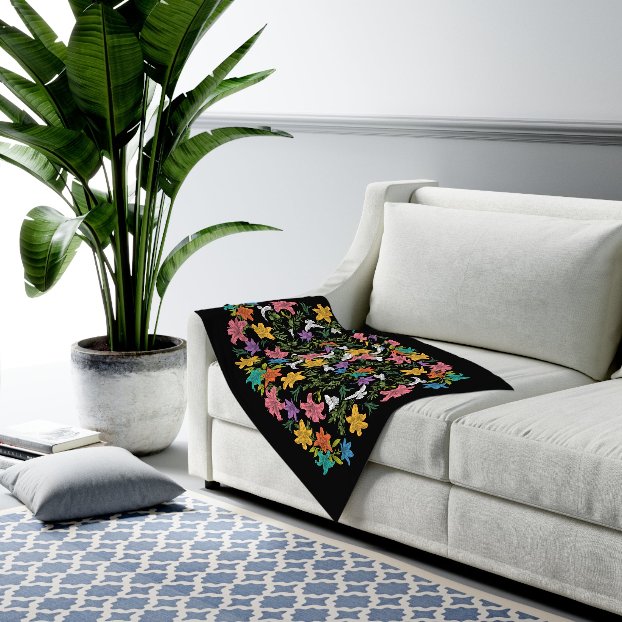 A black swaddle blanket draped over a transitional-styled sofa. The blanket has an all-over, multi-colored pattern of doves, flowers, and olive branches.