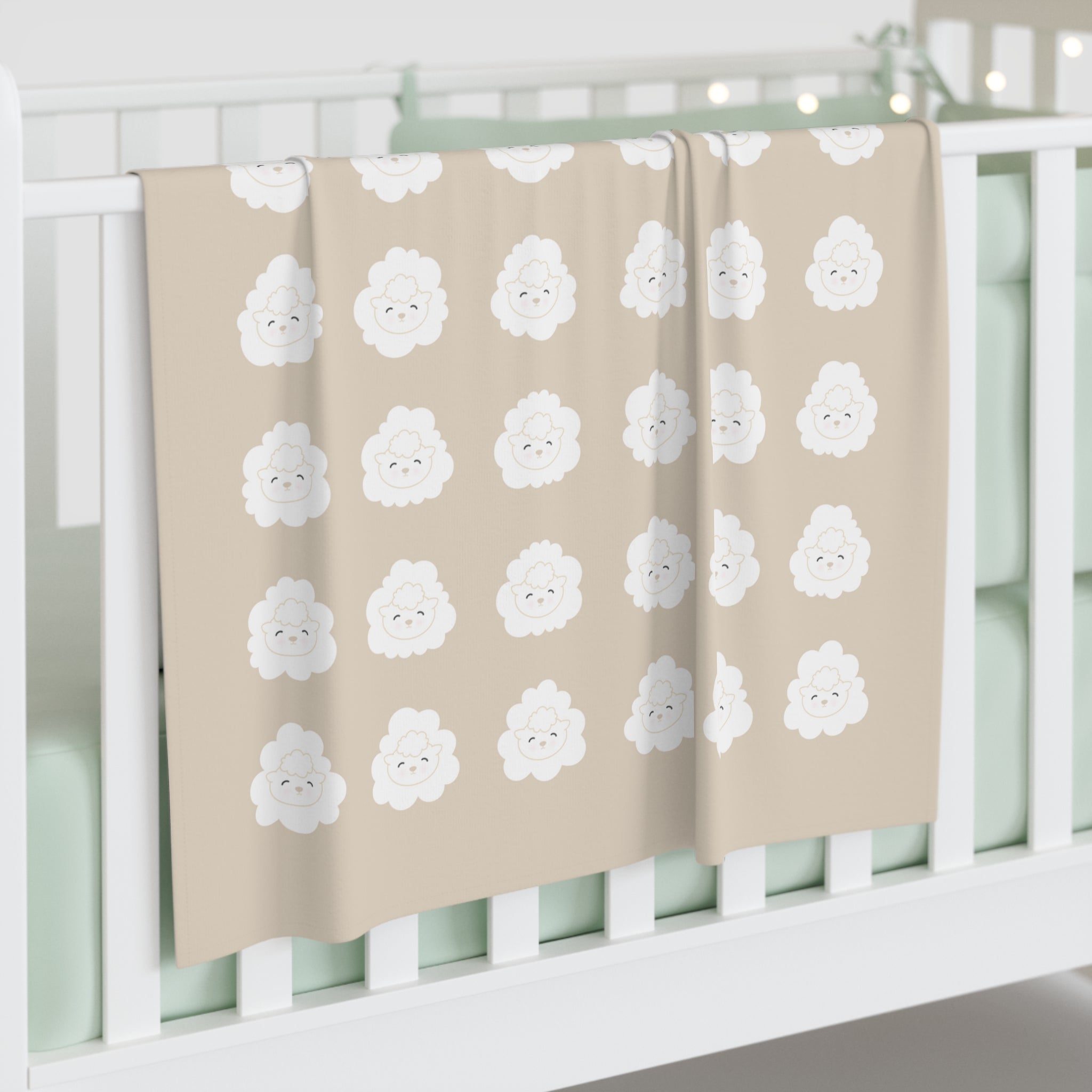 A beige swaddle blanket draped over the side of a crib. The blanket has an all-over, white, lamb-face pattern in even rows.