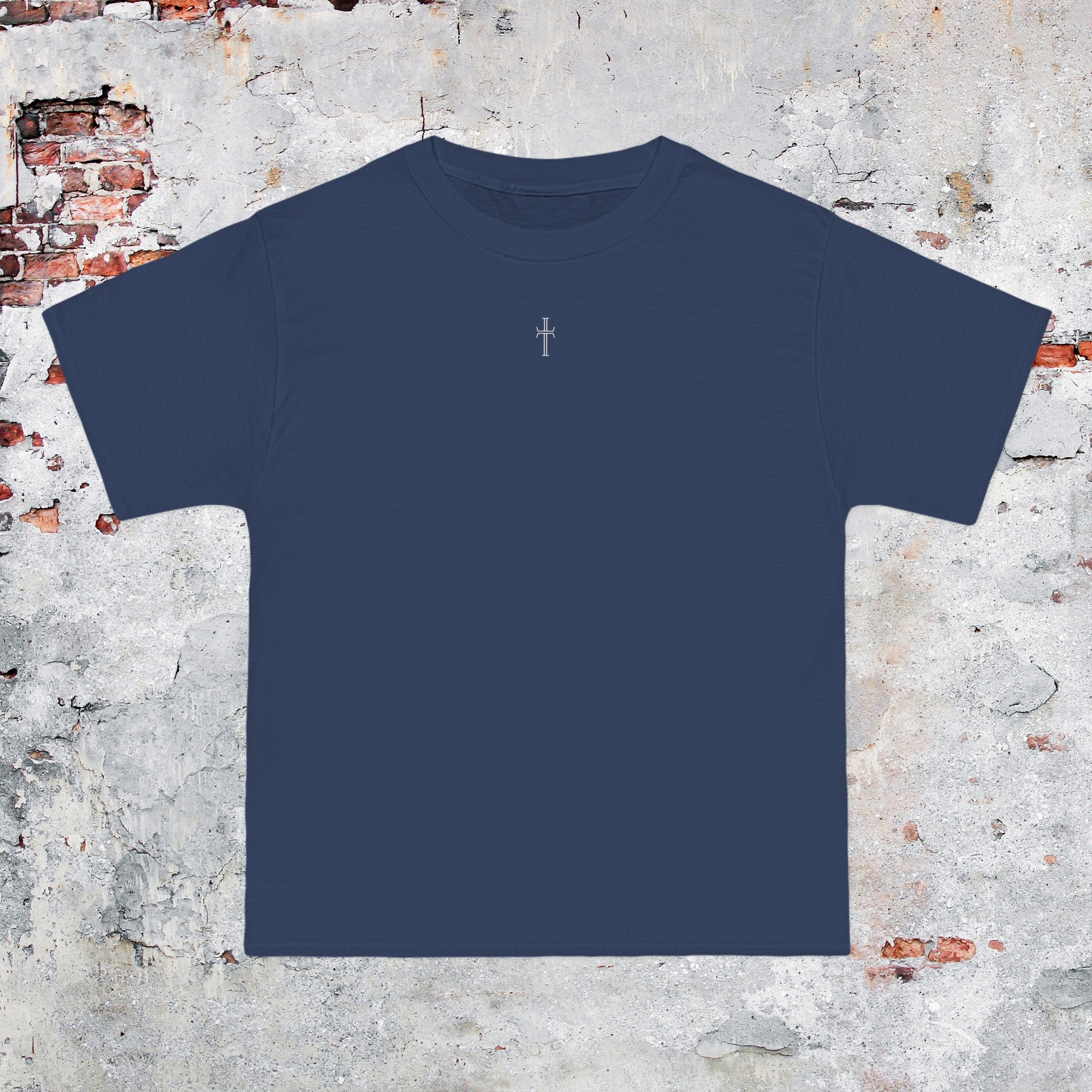 A denim blue crewneck tee shirt displays the front of the garment as it lays against a brick-patterned wall. A small white Purpose cross logo is printed at the top center. 