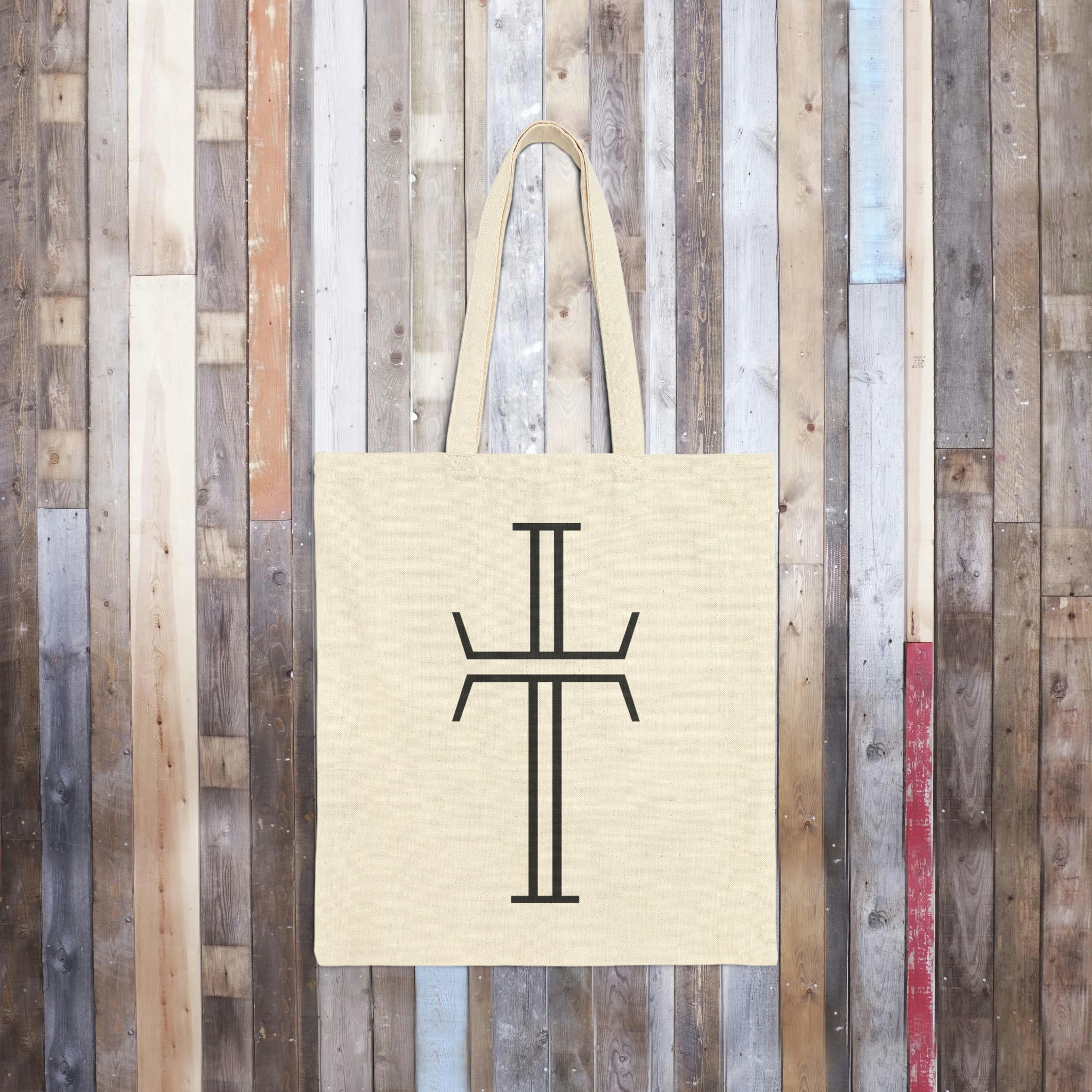 A natural canvas bag lays flat on a wood floor. The bag is rectangular with long looped handles and displays a black Purpose cross logo on the front.