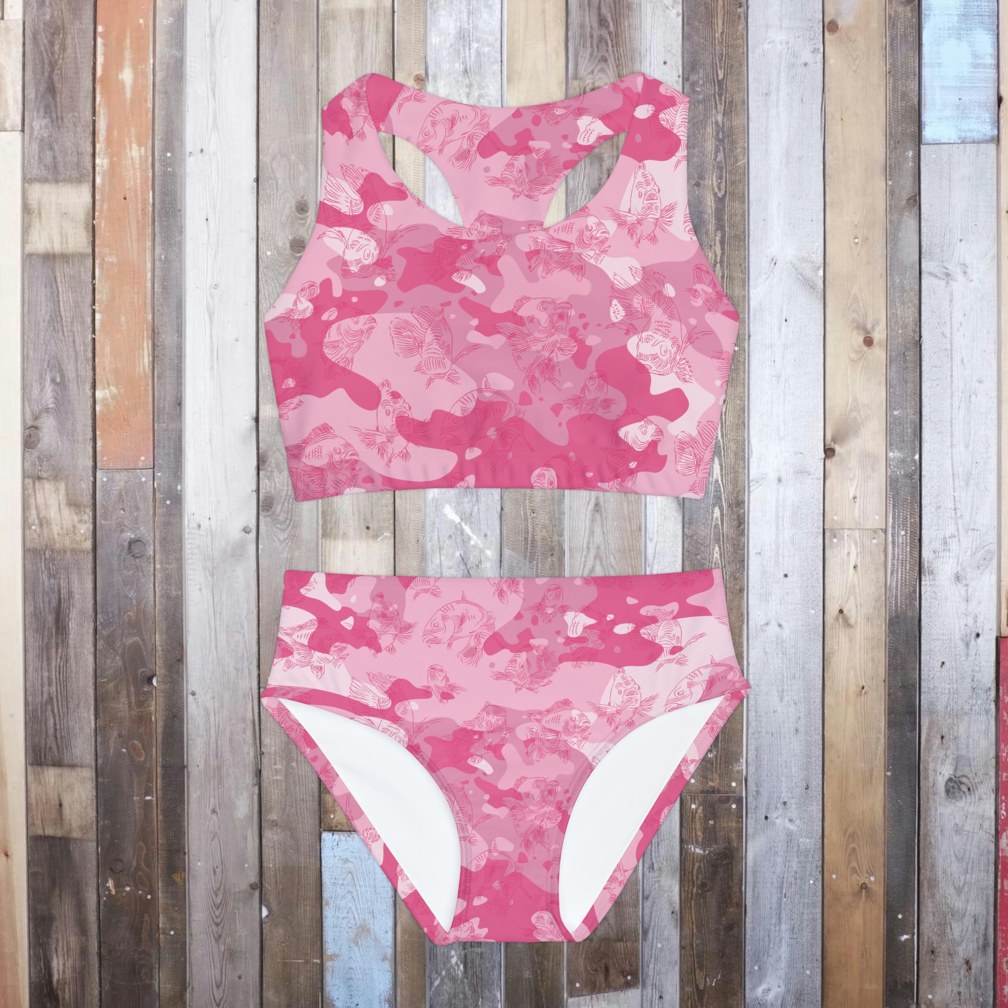 A two-piece tank-style swimsuit laying flat on a hardwood floor, in a pink-camo design with an all-over swimming carp pattern in dark pink. 