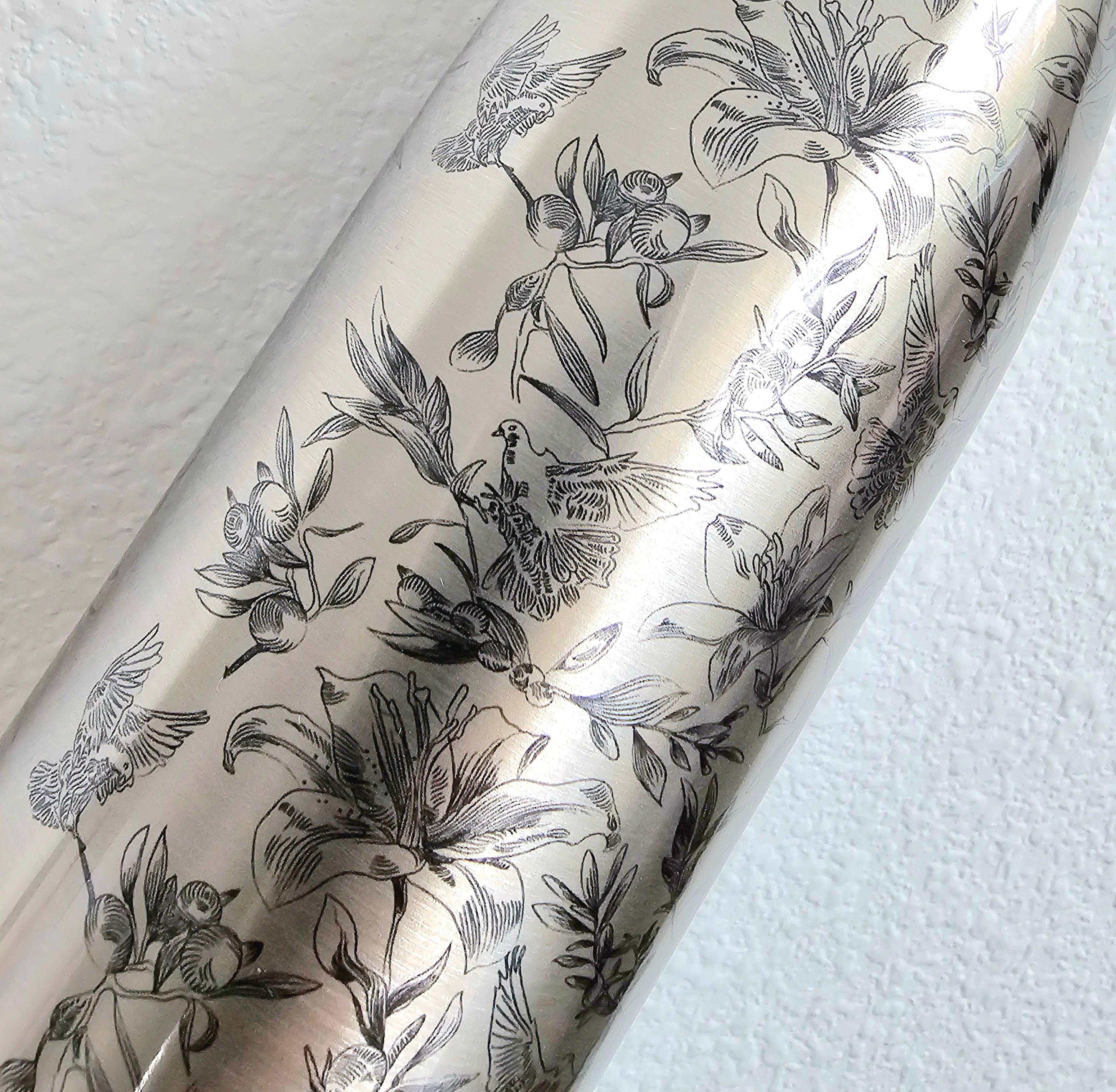 Close up of a stainless steel water pattern. It displays a black floral, olive leaf, and dove pattern against a white textured wall.