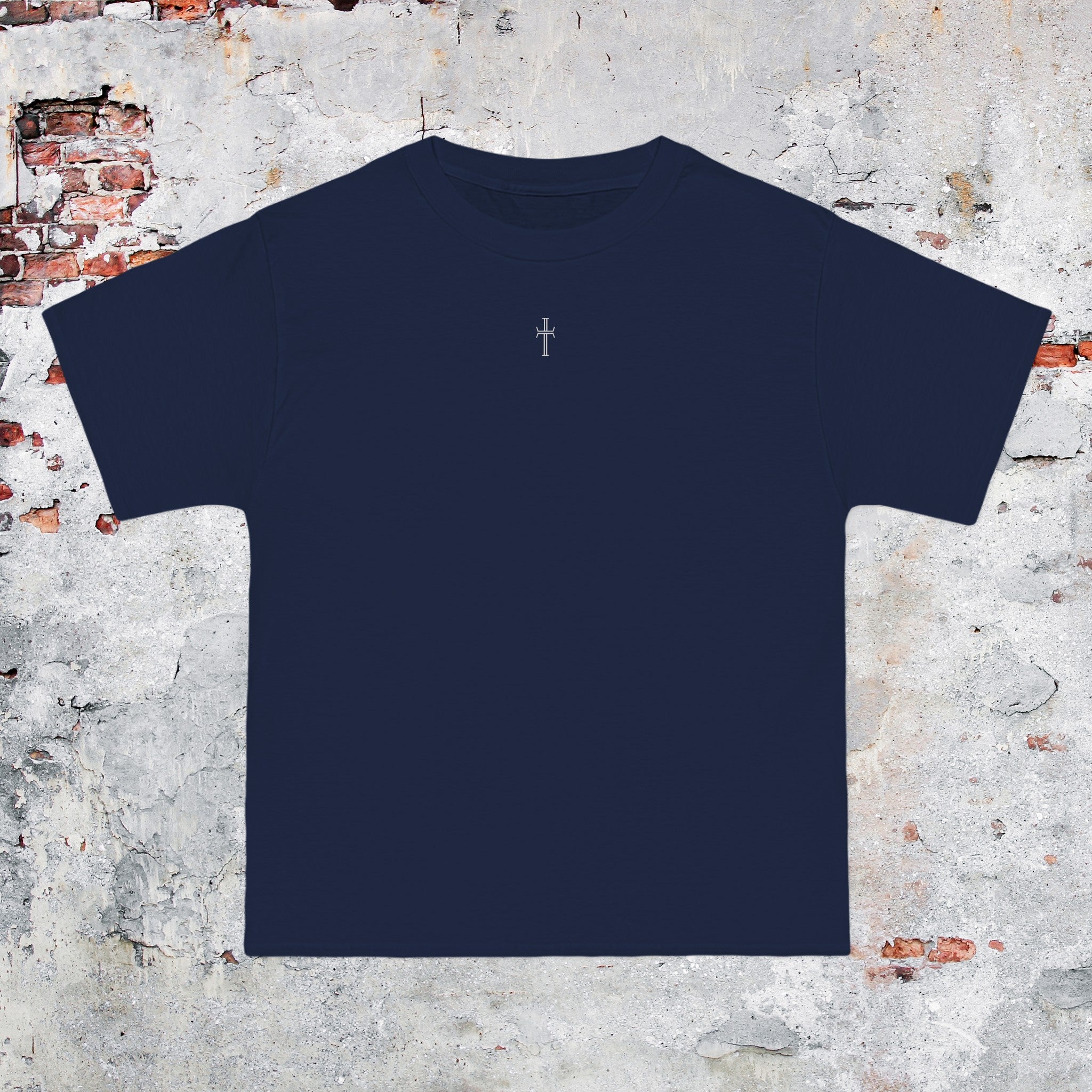 A navy crewneck tee shirt displays the front of the garment as it lays against a brick-patterned wall. A small white Purpose cross logo is printed at the top center. 