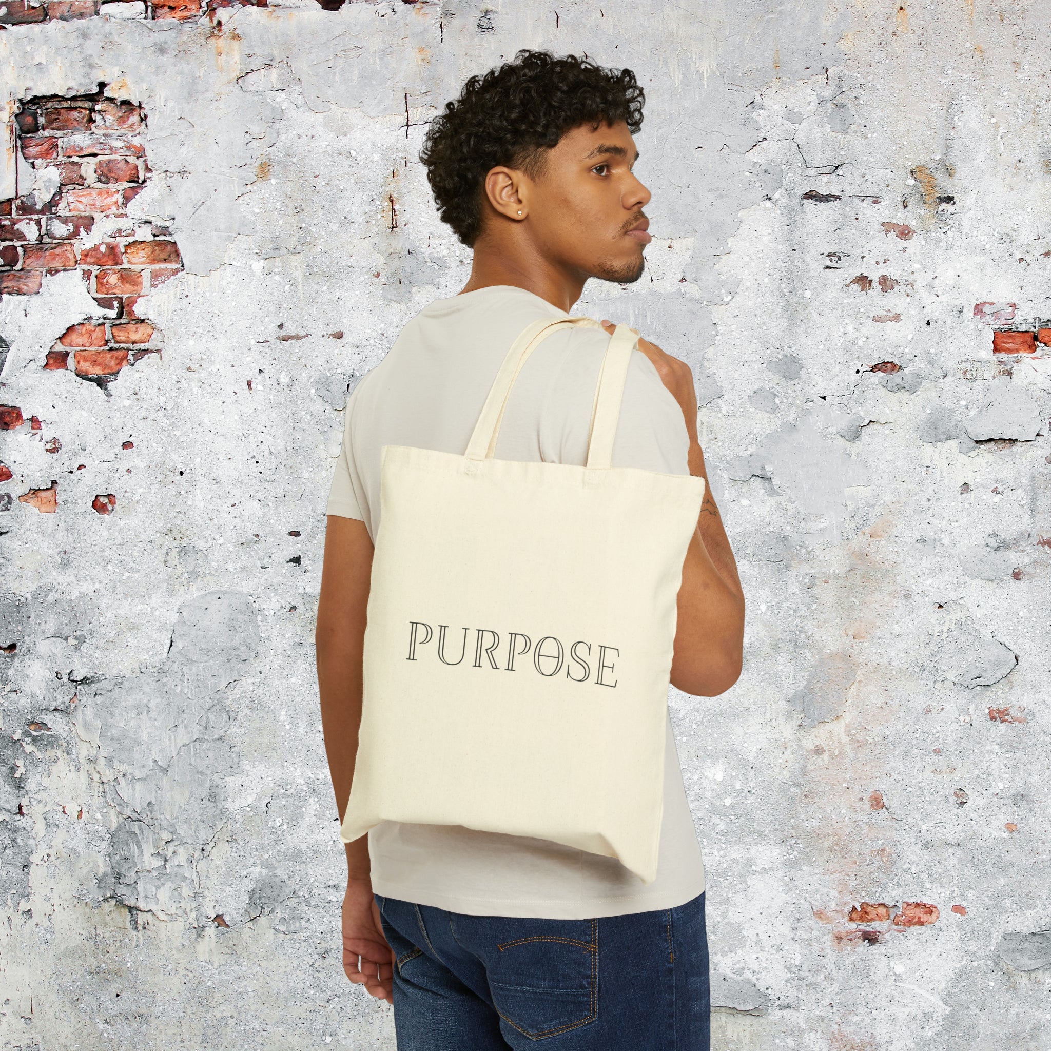 A male model in a tee shirt and jeans is slinging a tote bag with long double handles over the back of his shoulder. The natural canvas bag has the black Purpose word logo on the back of the bag.