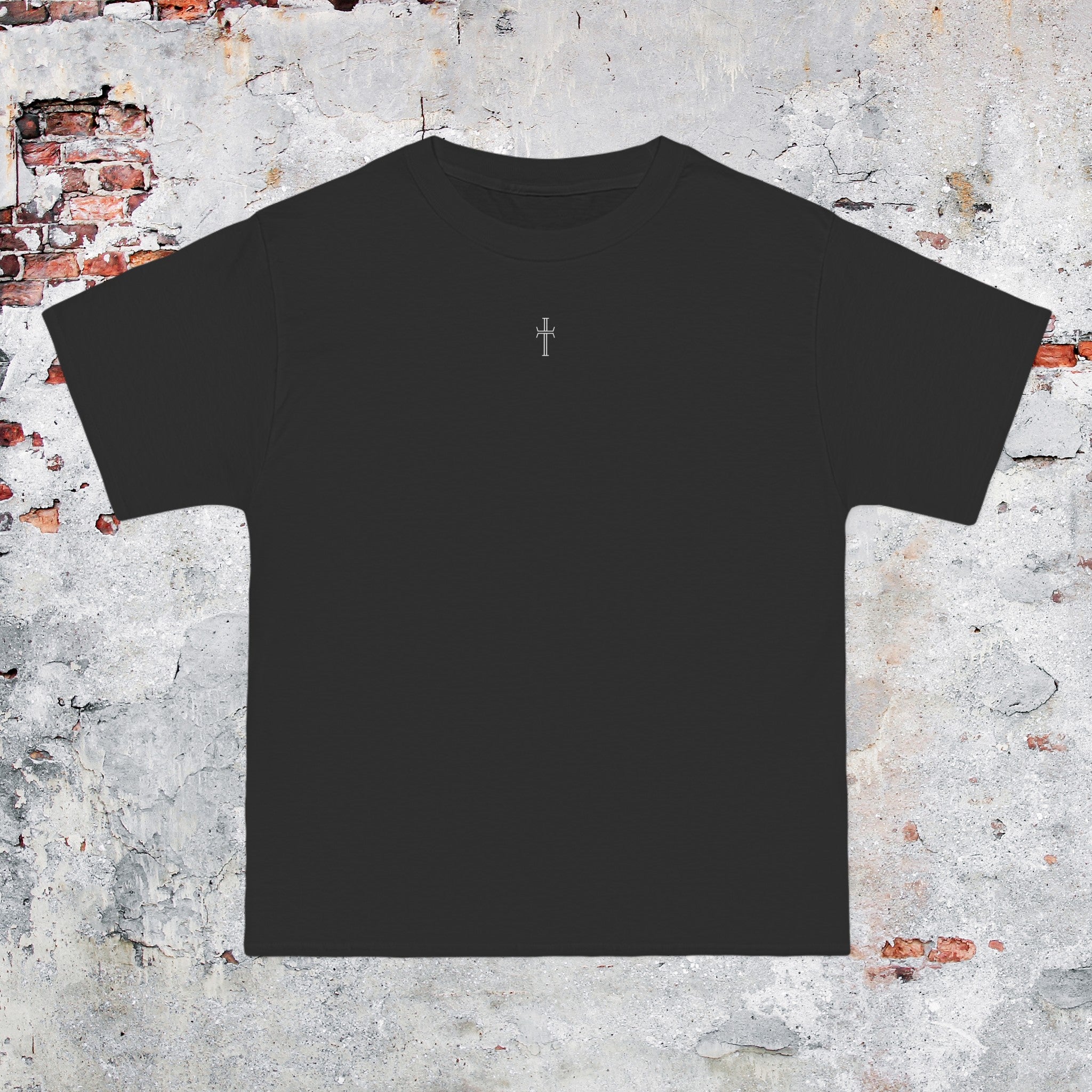 A black crewneck tee shirt displays the front of the garment as it lays against a brick-patterned wall. A small white Purpose cross logo is printed at the top center. 