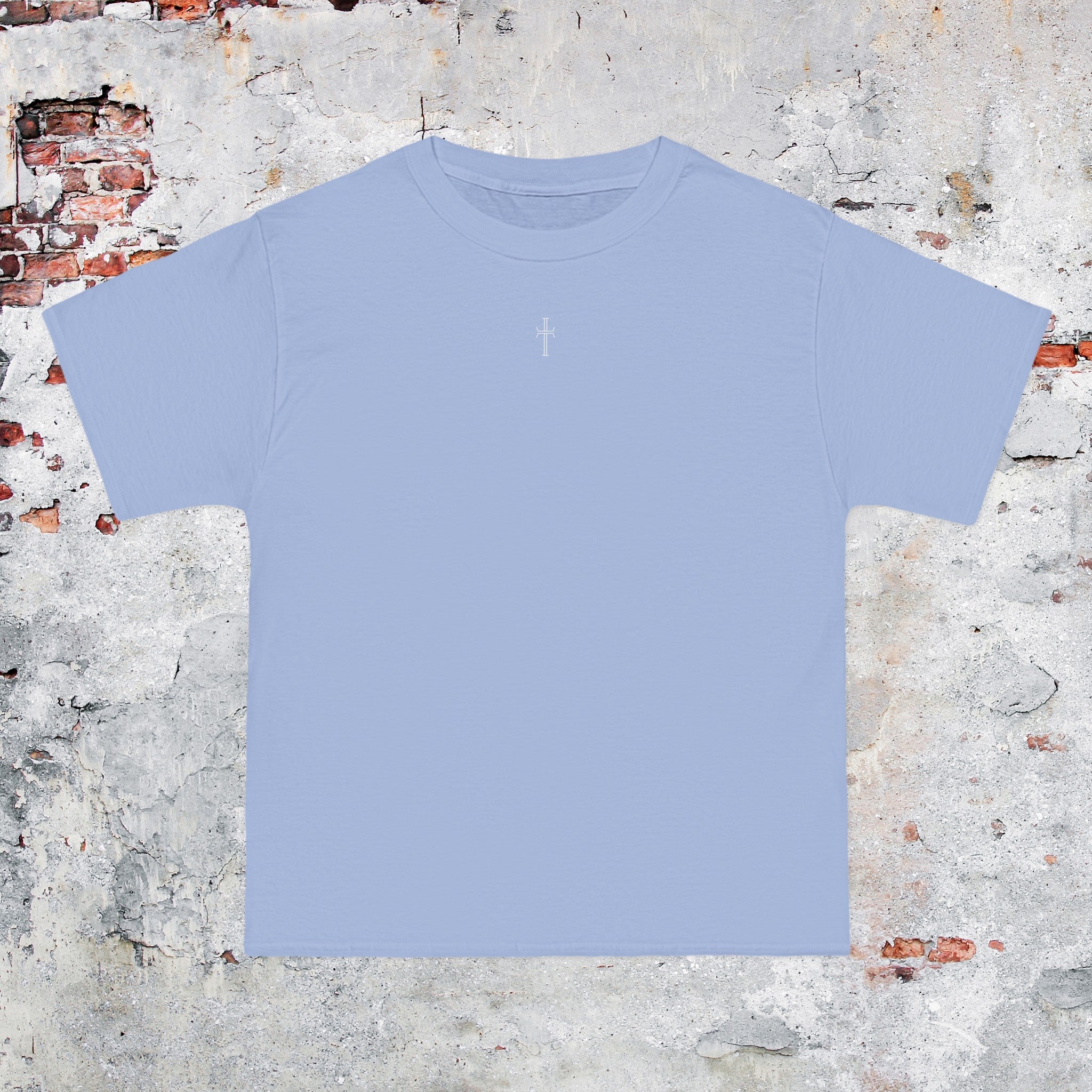 A light blue crewneck tee shirt displays the front of the garment as it lays against a brick-patterned wall. A small white Purpose cross logo is printed at the top center. 