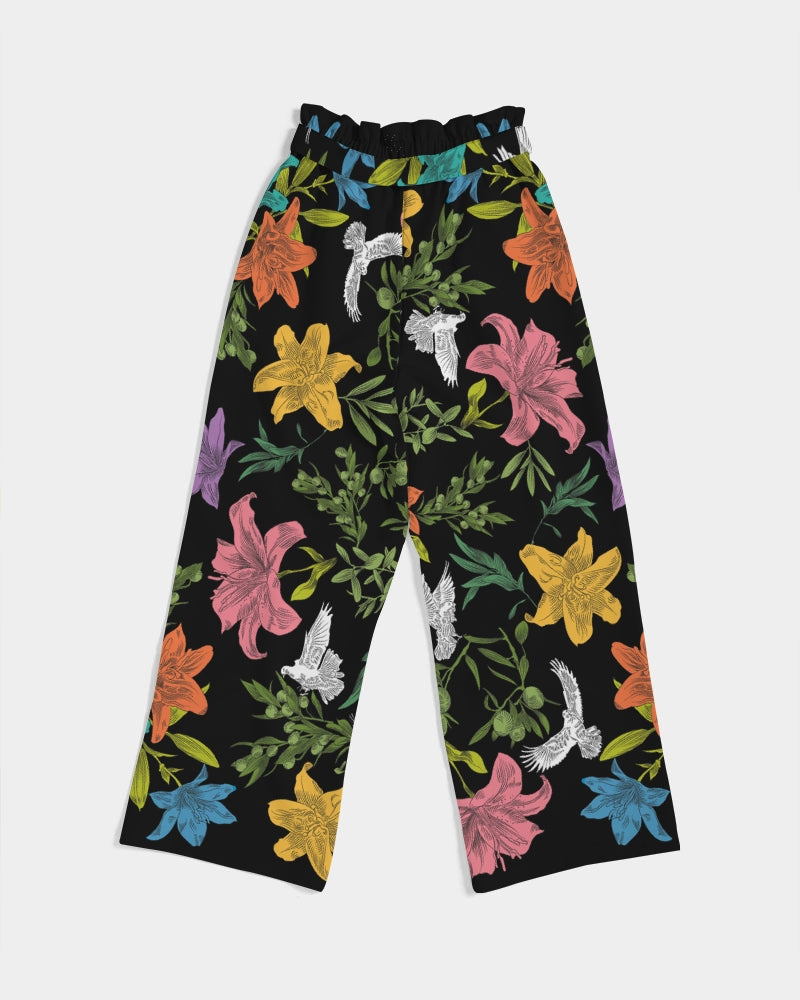 Back view of high-waisted, wide-leg black pants with a bold, multi-colored pattern of lilies, white doves, and olive branches in a large, all-over print with self-tie belt and elastic waist, laying flat against a white background.