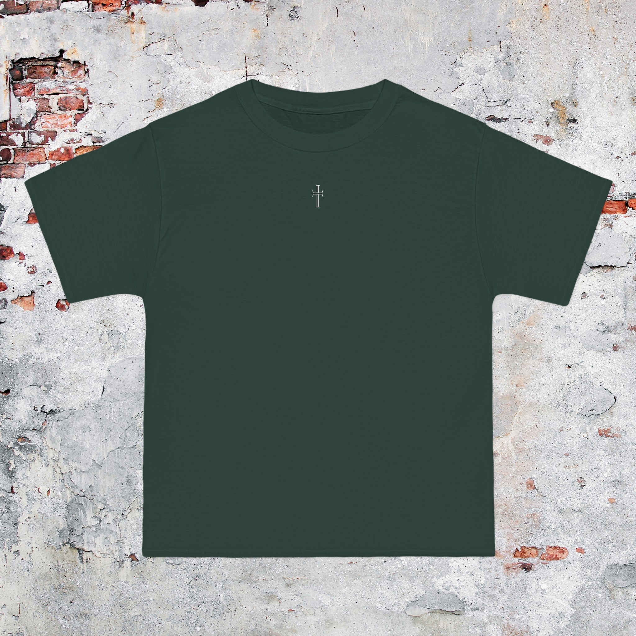 A forest green crewneck tee shirt displays the front of the garment as it lays against a brick-patterned wall. A small white Purpose cross logo is printed at the top center. 