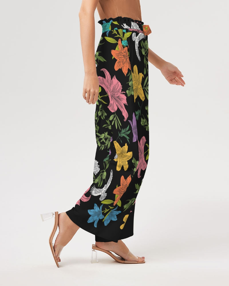 Right view of bottom half of female model wearing high-waisted, wide-leg black pants with a bold, multi-colored pattern of lilies, white doves, and olive branches in a large, all-over print with self-tie belt and elastic waist.